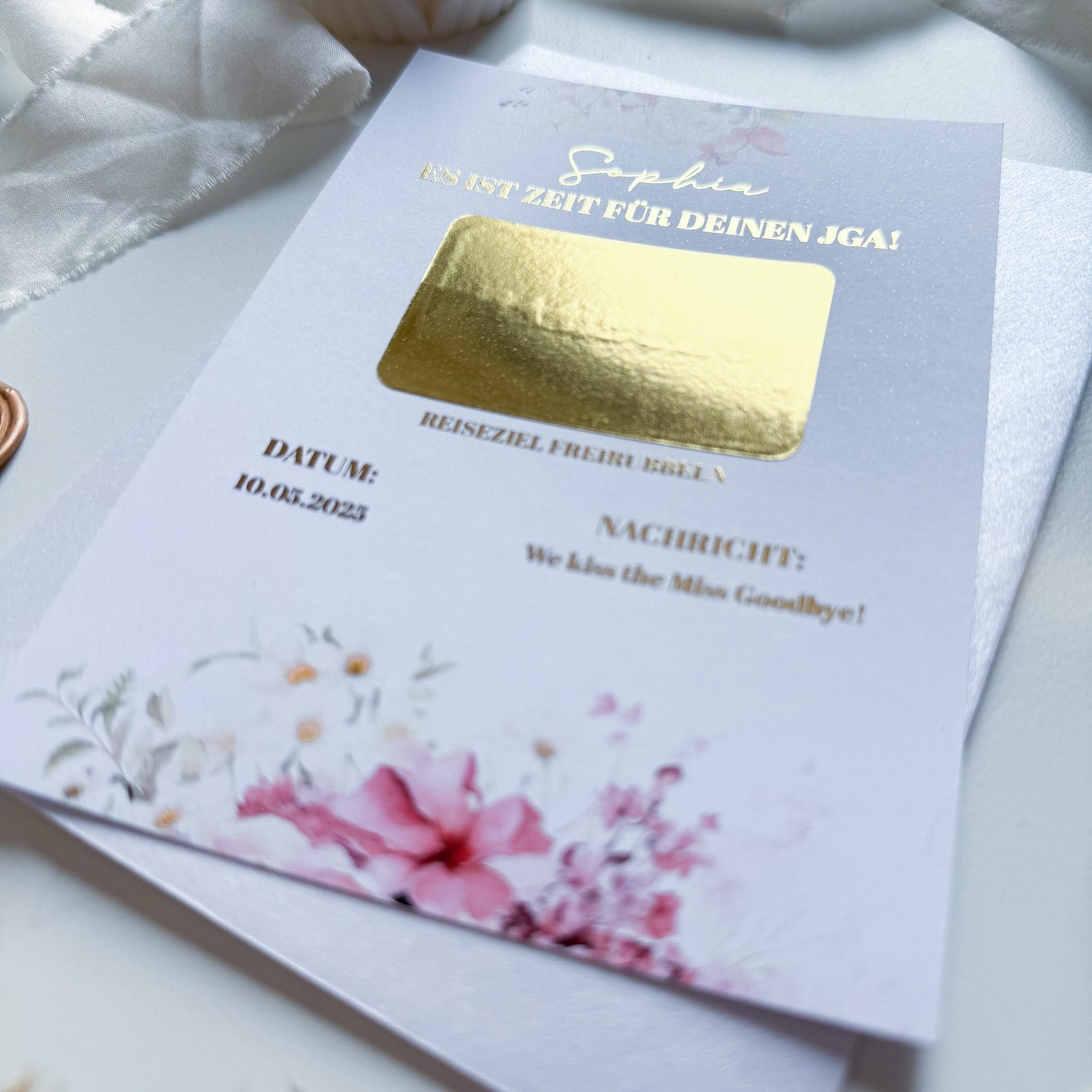 Eine Nahaufnahme der Funkenschauer-Rubelkarte „Einladung zum JGA - JGA Gutschein“ zeigt das A6-Design Pastelltraum mit Goldfolie, handschriftlicher und gedruckter deutscher Schrift auf weißem Stoff-Hintergrund.