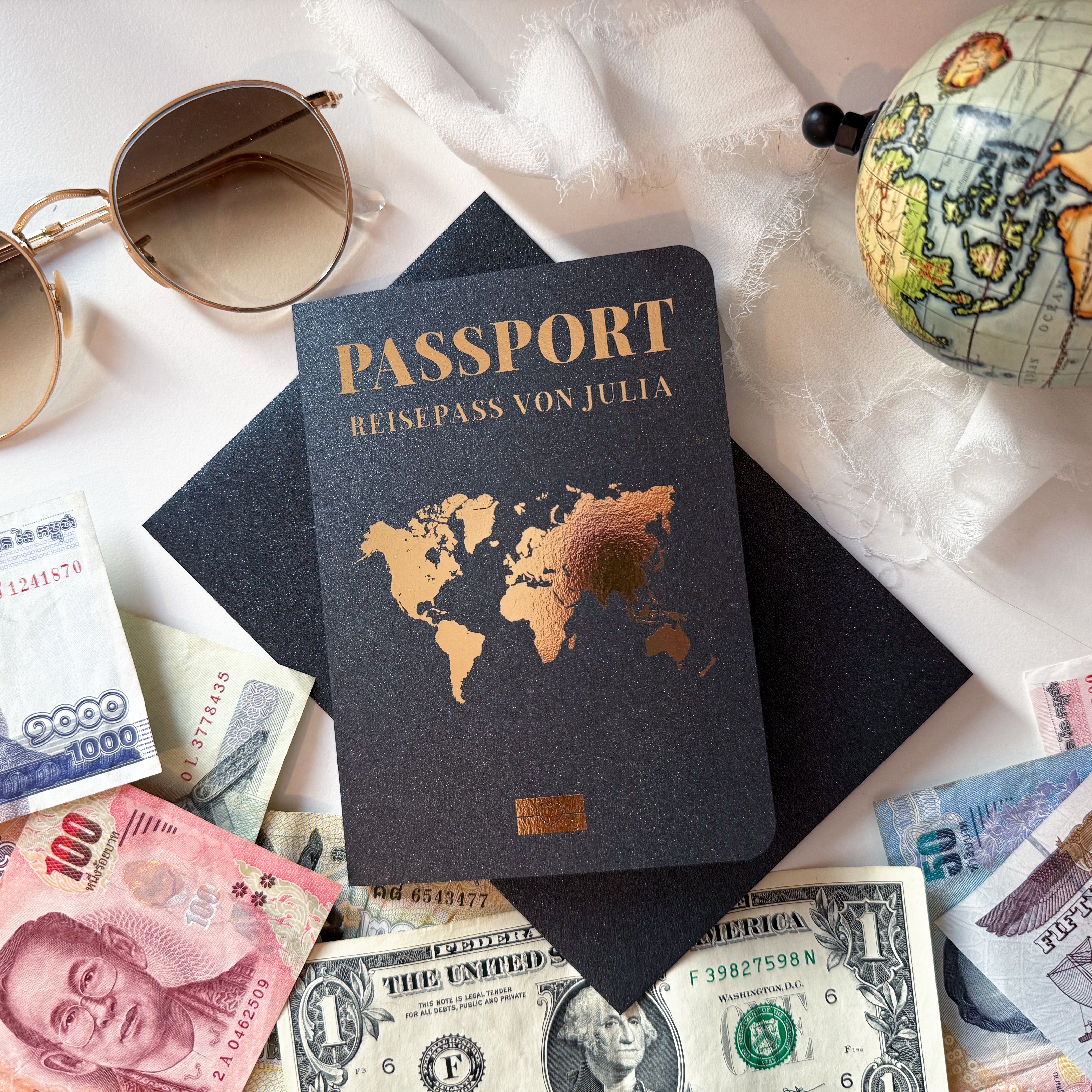 Ein Flatlay zum Thema Reisen zeigt funkenschauers „Urlaub verschenken“, einen Reisepass mit Weltkartencover, internationaler Währung, Sonnenbrille, Globus und schwarzem Umschlag. Der weiße Stoffhintergrund hebt diesen personalisierten Reisegutschein hervor – ideal als einzigartiges Geburtstagsgeschenk oder Reisegutschein-Idee.