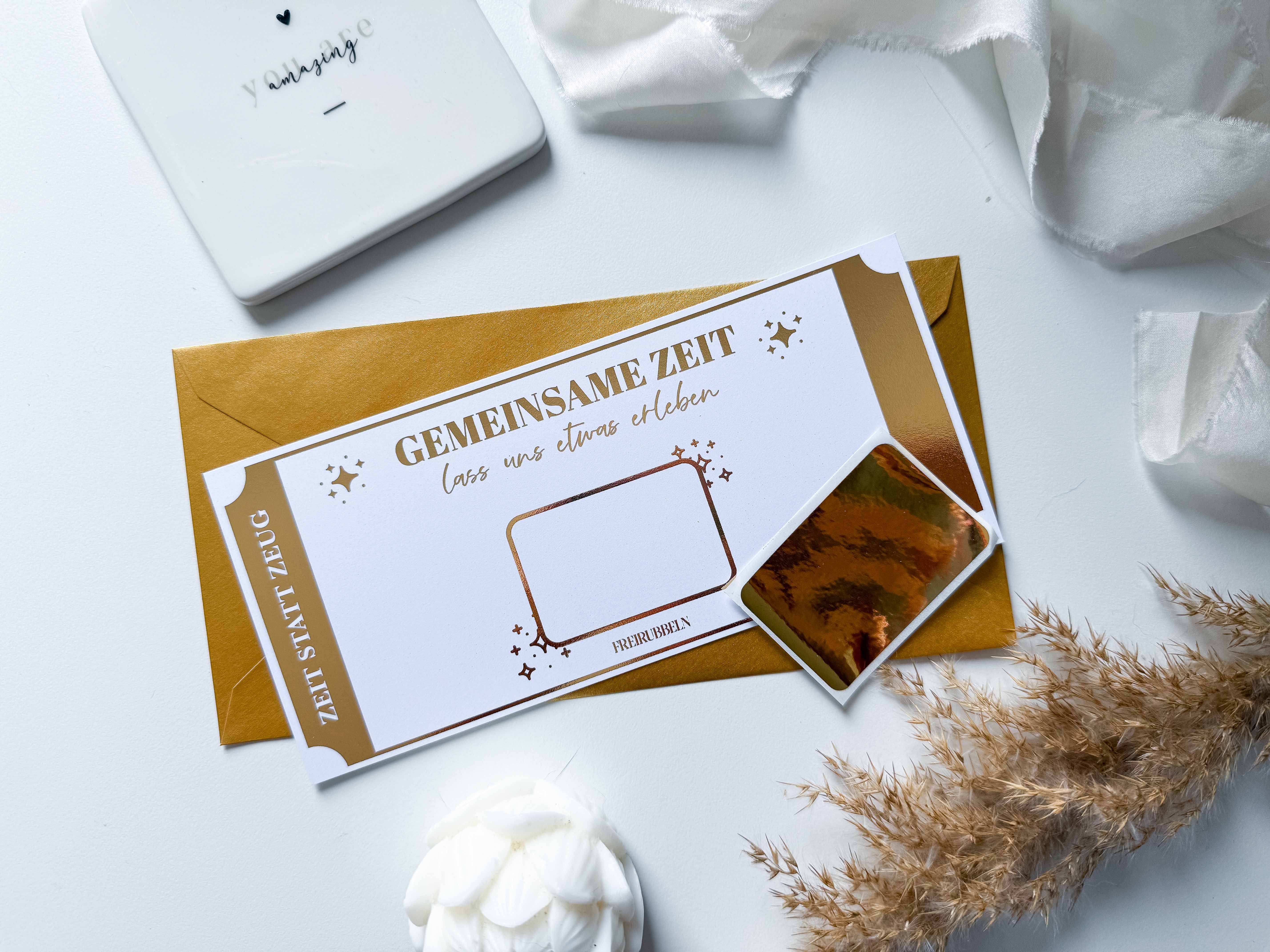 Ein deutscher „Zeit statt Zeug“-Gutschein von funkenschauer mit der Aufschrift „GEMEINSAME ZEIT“ liegt auf einem braunen Umschlag und wird wunderschön neben einem beigefarbenen Stoff, einem Untersetzer mit abstraktem Muster, einer weißen Rose und getrocknetem Gras präsentiert – eine elegante, handgefertigte Präsentation.