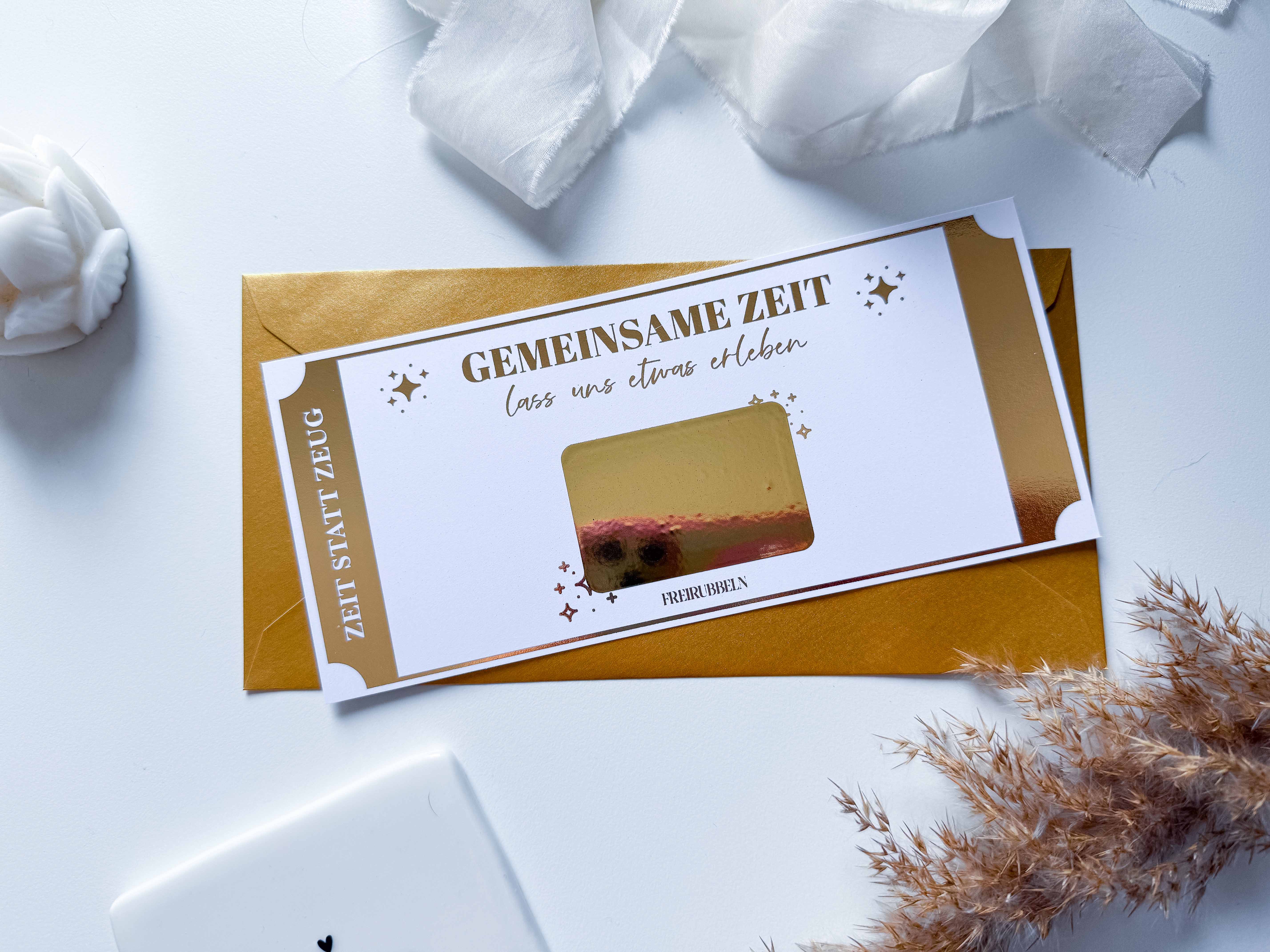 Ein handgefertigter „Zeit statt Zeug Gutschein“ von Funkenschauer besteht aus einer individualisierbaren Geschenkkarte, die in einem eleganten Umschlag auf einer weißen Oberfläche verpackt ist. Auf dem Ticket steht „Gemeinsame Zeit, lass uns etwas erleben“ und ist zur Personalisierung mit weißem Stoff und getrockneten Pflanzen verziert.