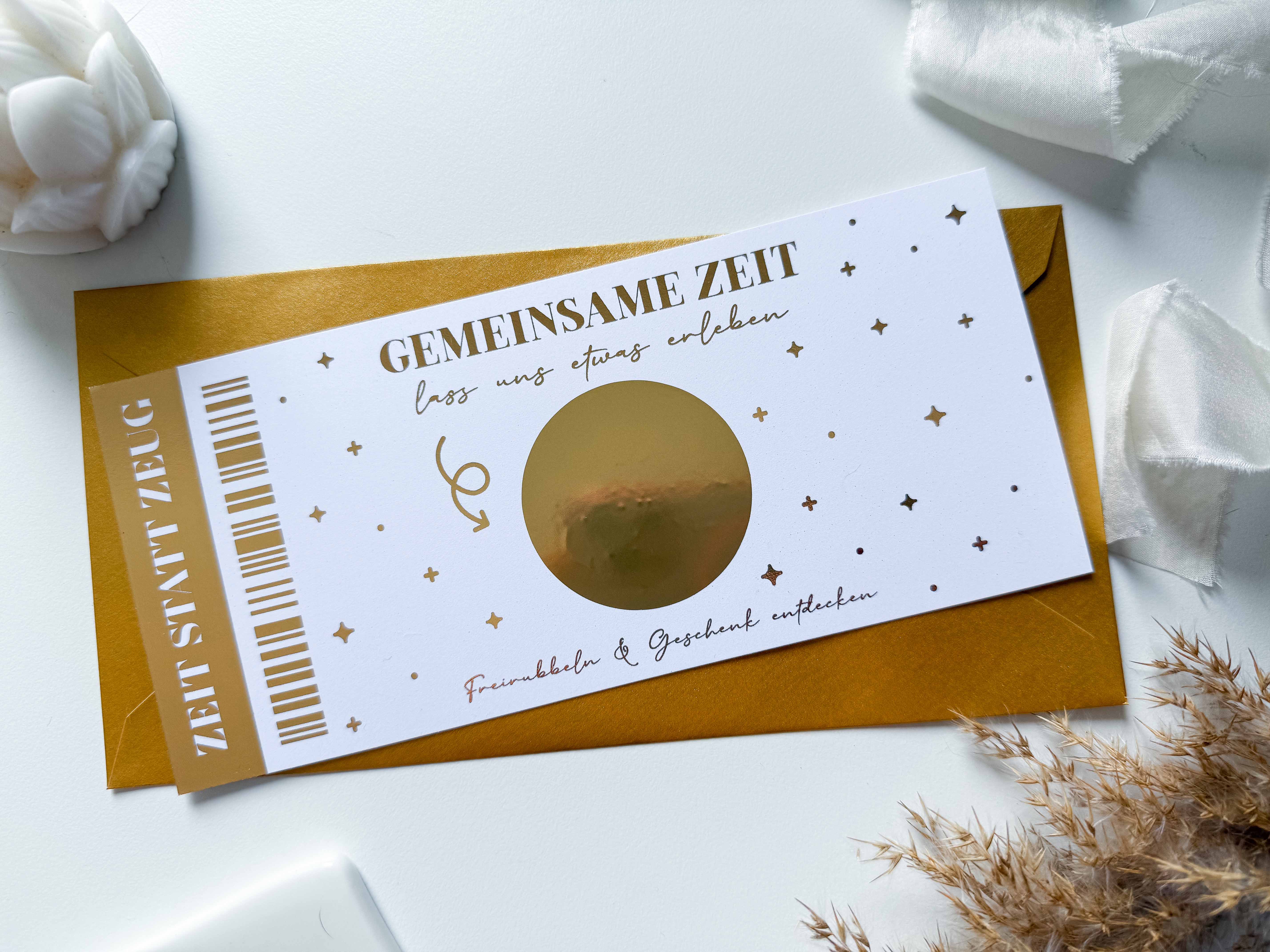 Auf einem Tisch liegt eine Karte von Funkenschauer mit goldenen und weißen Motiven, in deren Mitte „Gemeinsame Zeit“ und eine goldene Rubbelkarte stehen. Es ist mit einem goldenen Umschlag versehen und von sanften Dekorationen umgeben – ein herrlicher Gutschein Zeit statt Zeug personalisiert.