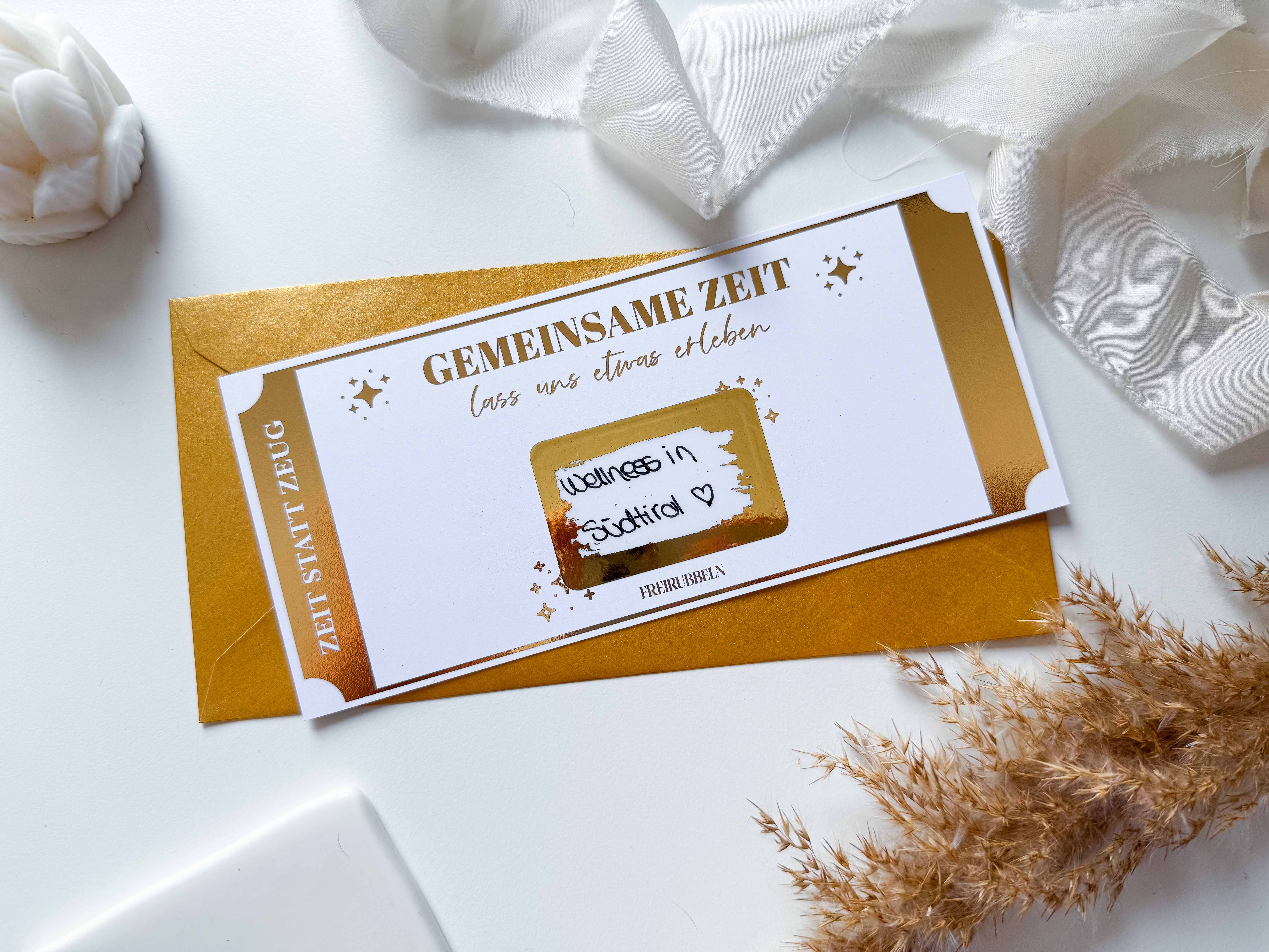 Ein gold-weißer „Zeit statt Zeug“-Geschenkgutschein von funkenschauer, verziert mit deutschem Text und Sternen, liegt neben einem passenden Umschlag auf einer Oberfläche mit getrocknetem Pampasgras und hauchdünnem weißen Stoff.