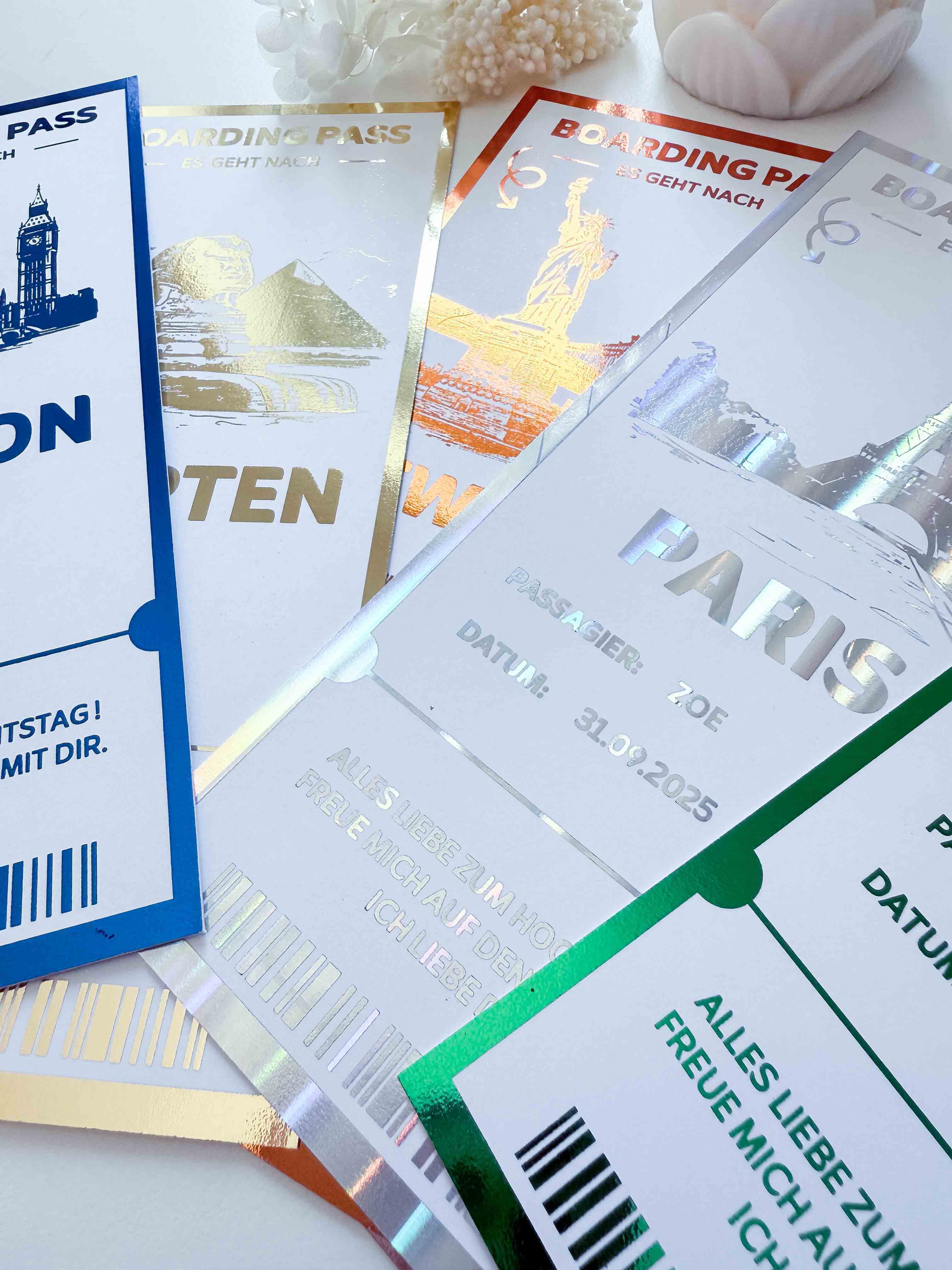 Eine Nahaufnahme der personalisierten Reisegutschein-Karten von funkenschauer in schillernden Farben, stilvoll im Boarding-Pass-Design mit Zielen wie Paris und New York-arrangiert fächerförmig auf Weiß mit Trockenblumen, ideal als Geschenk für Reisende.