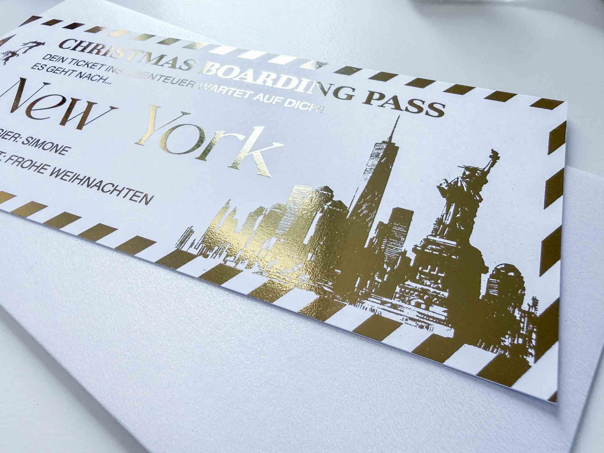 Eine Nahaufnahme eines funkenschauer Reisegutscheins Weihnachtsgutschein, personalisiert mit New York-Skyline und Freiheitsstatue in Goldfolienprägung, auf einem weißen Umschlag - perfekte Geschenkidee zu Weihnachten auf Deutsch und Englisch.
