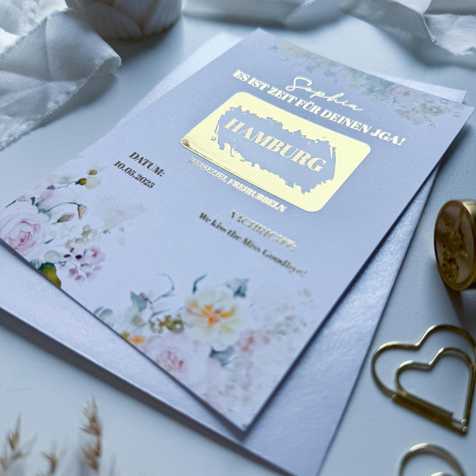 Eine Nahaufnahme des funkenschauer JGA Gutschein – Rubbelkarte A6 Vintage Rosé, verziert mit goldener Schrift „Hamburg“, auf rosegoldenem Perlmuttpapier. Umgeben von Bändern, Herz-Paperclip und Ring – ideal als Reisegutschein zum Junggesellinnenabschied.