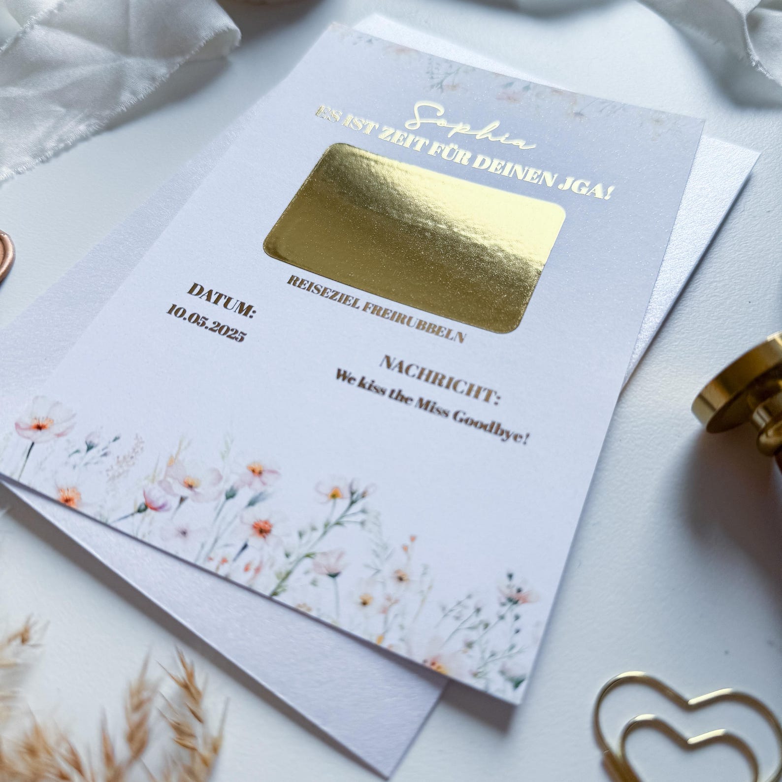 Eine Nahaufnahme des funkenschauer JGA Gutscheins – personalisierte A6 Rubbelkarte Gänseblüten mit Goldfolie, deutschem Text und Rubbelbereich „Bride to Be“, umgeben von weißem Band und goldener Büroklammer.