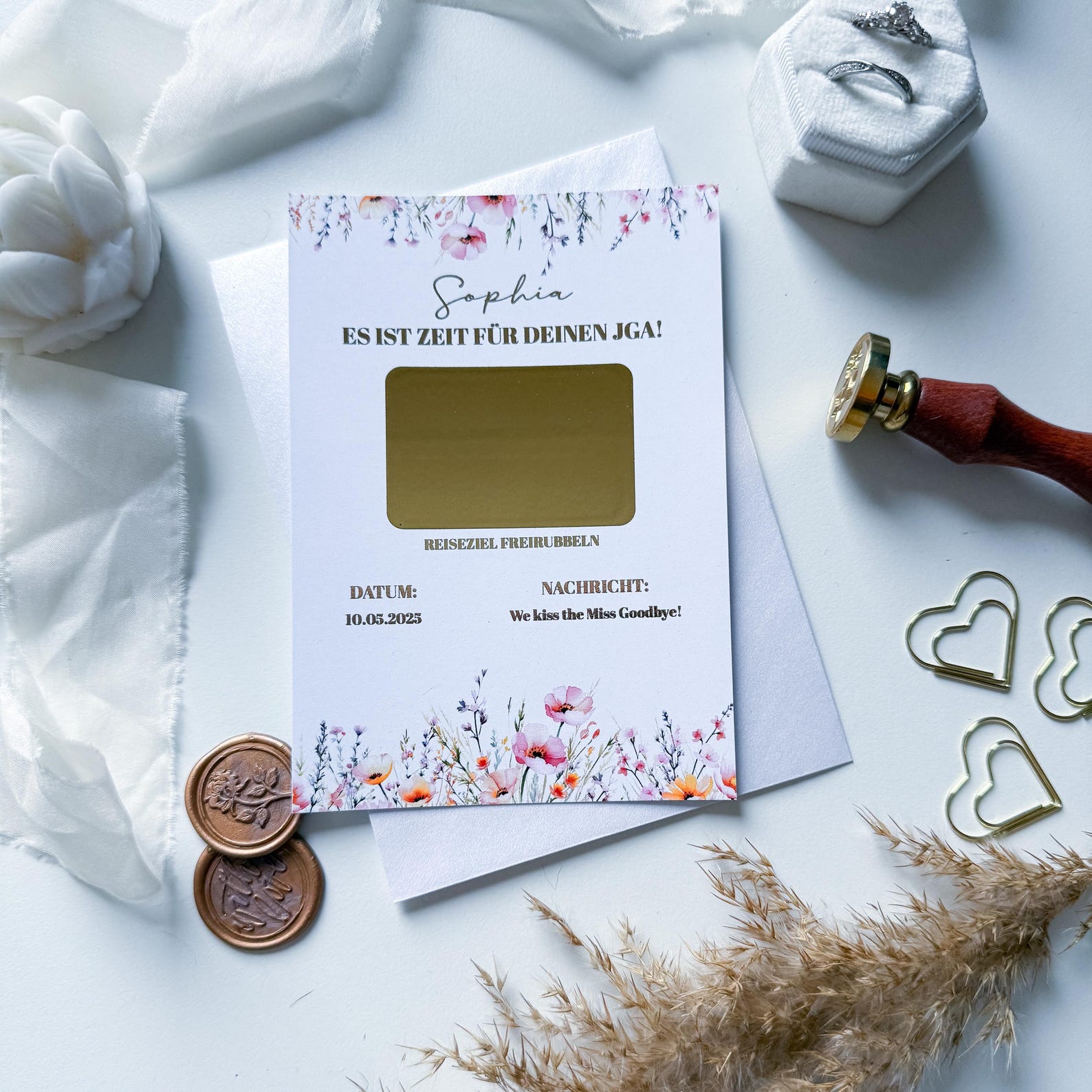 Die Rubbelkarte „JGA Gutschein – Wiesenglück“ von Funkenschauer zeigt florales Design, goldene Rubbelfläche und persönlichen Einladungstext zum JGA auf Deutsch. Dekoriert mit Münzen, Ring-Etui, Wachssiegel und Herzen.