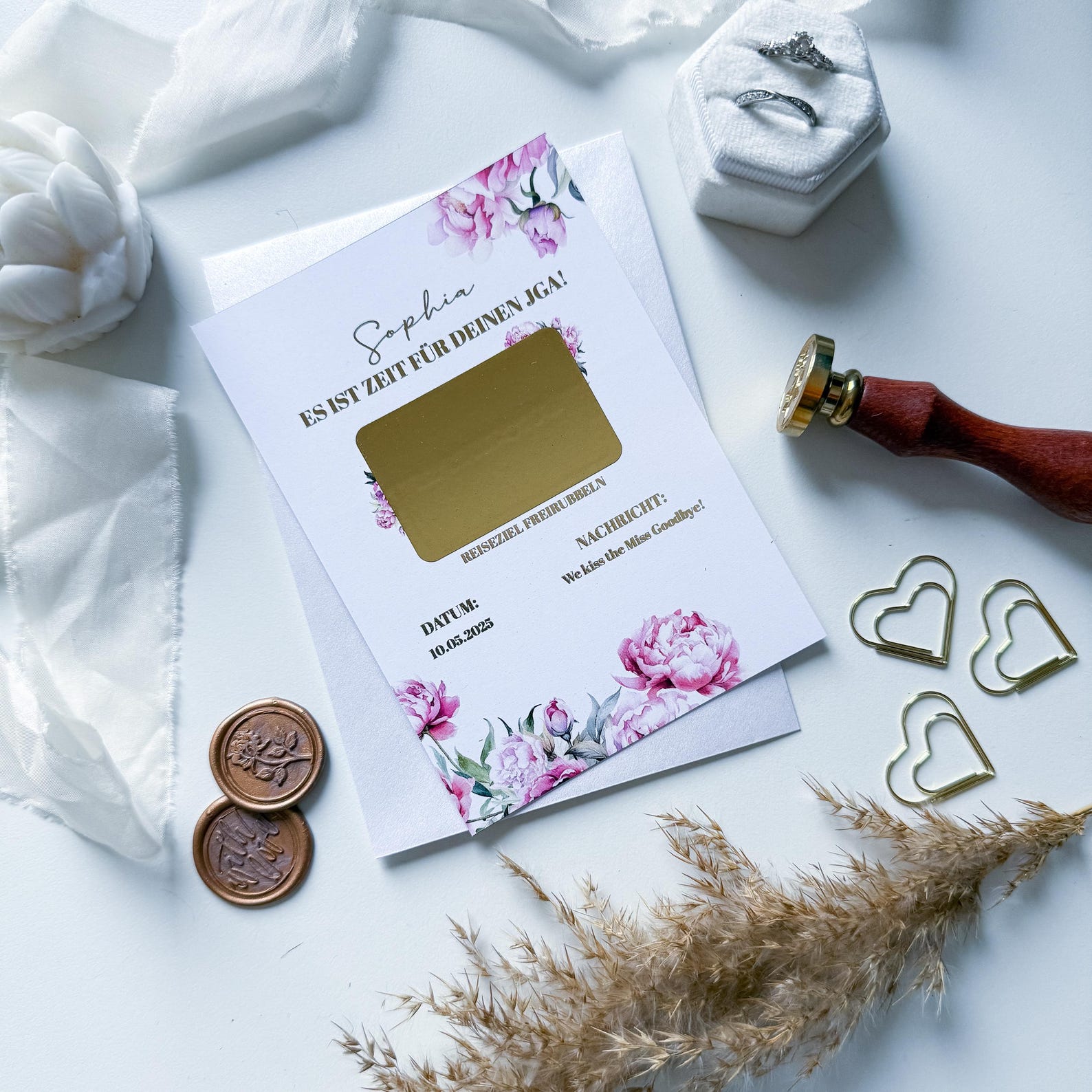 Der funkenschauer JGA Gutschein – Reisegutschein verschenken ist eine florale A6 Rubbelkarte „Pfingstrosenzauber“, abgebildet mit Eheringen in einer weißen Schachtel, einem Wachssiegelstempel, goldenen Herz-Büroklammern, Münzen und getrocknetem Gras auf weißem Untergrund.