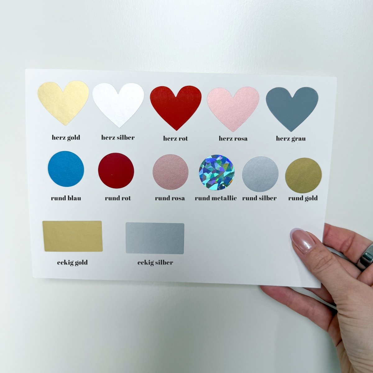 Eine Hand hält eine Funkenschauer-Karte mit metallischen Gold-, Silber-, Rot-, Blau-, Rosa-, Grau- und holografischen Aufklebern in Herzen, Kreisen und Rechtecken – jeweils beschriftet auf Deutsch. Perfekt zum Verschenken von Reisegutscheinen & Geschenk für Reiseliebhaber.