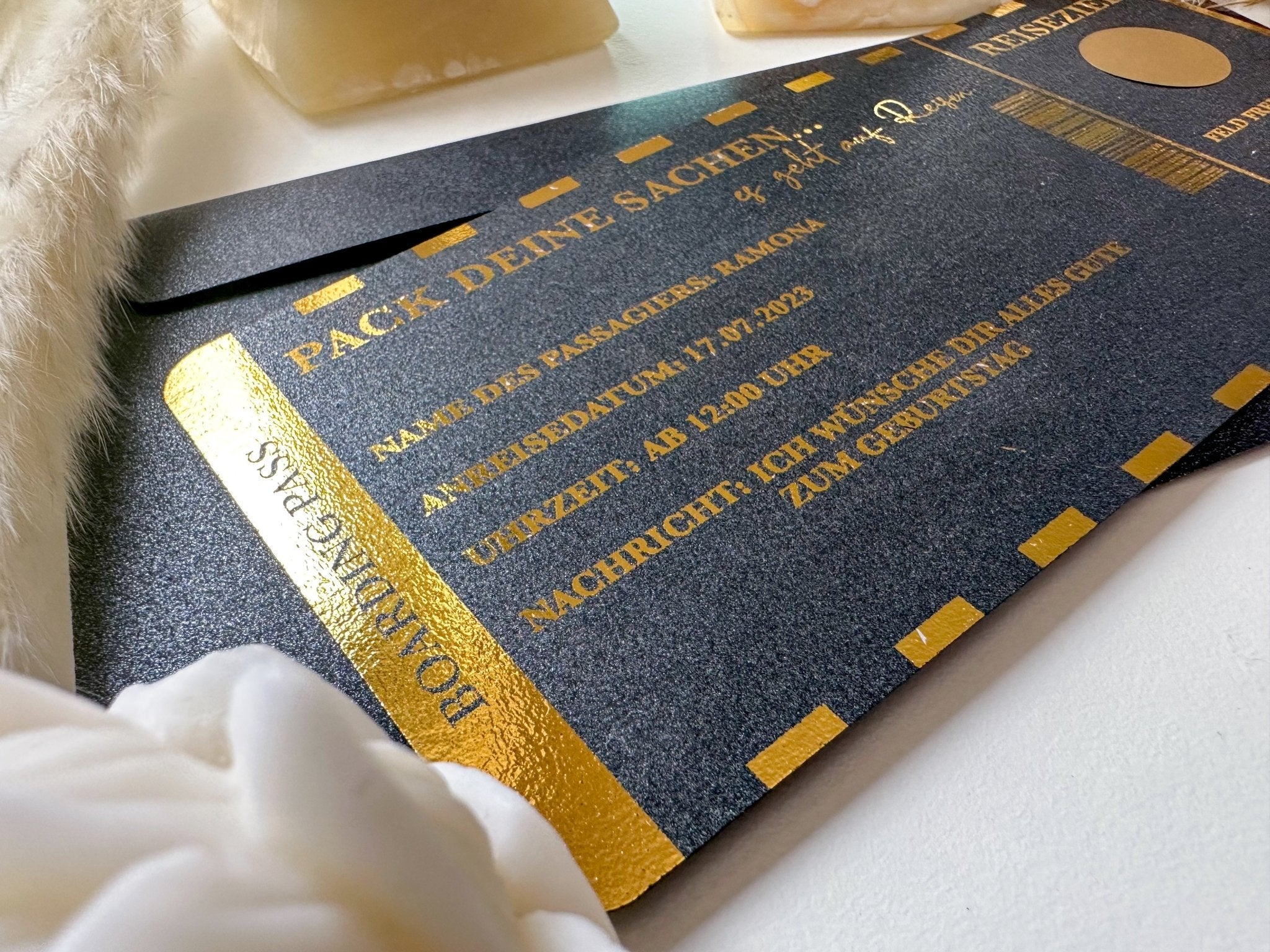 Eine Nahaufnahme der Funkenschauer-Karte „Reisegutschein verschenken • Gutschein Personalisiert • Geschenk für Reiseliebhaber“, schwarz mit goldglänzendem Schriftzug, gestaltet wie ein Reisedokument, auf einer weißen Fläche mit Dekoartikeln liegend.