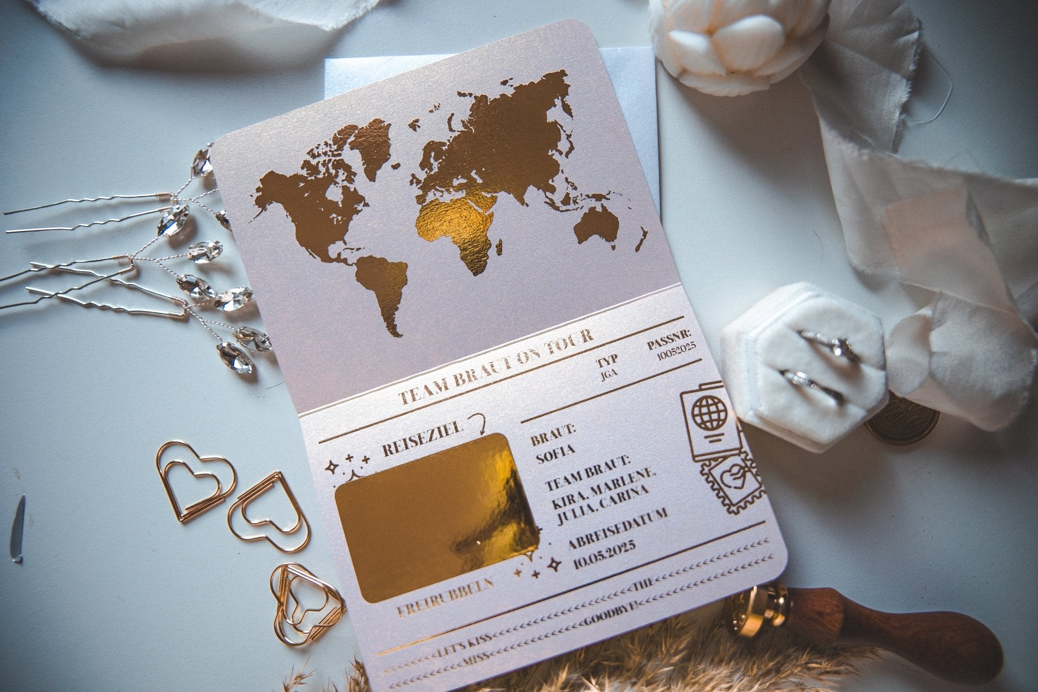 Die Junggesellinnenabschied Ideen Einladung von funkenschauer besticht durch ein Weltkarten-Design mit eleganten Goldfolien-Akzenten und herzförmigen Büroklammern. Daneben finden Sie eine Ringbox aus Samt und weiße Blumen – perfekt für kreative Partyideen.
