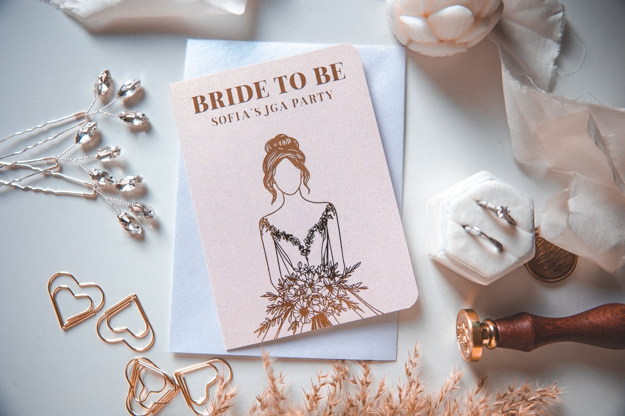 Die Einladungskarte „Junggesellinnenabschied Ideen“ von funkenschauer, bedruckt mit „Bride To Be“ und „Einladung JGA“, ist umgeben von Herzklammern, Diamantschmuck, einem Wachssiegel und einer Ringschachtel auf Stoff.