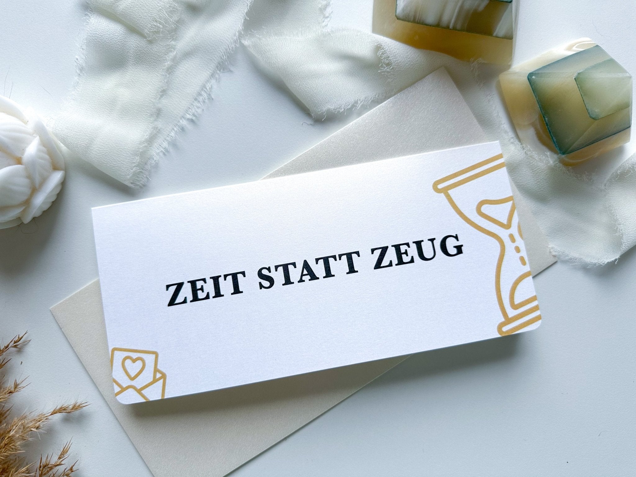 Eine personalisierte Rubbelkarte „Gutschein Zeit statt Zeug“ von funkenschauer, erhältlich in 17 Farben, mit kräftigem schwarzen Text auf weißer Karte, wird von einer weißen Kerze, weichen Stoffen, dekorativen Schachteln und getrockneten Blumen auf einer hellen Oberfläche begleitet.
