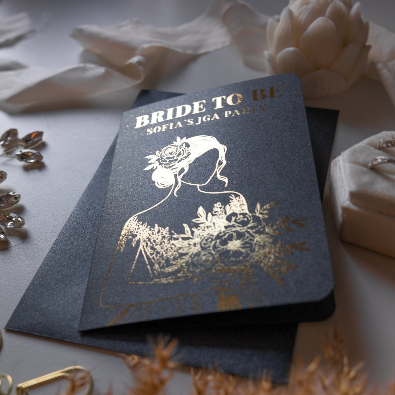 Die schwarze Karte mit Goldfolien-Design von funkenschauer zeigt eine Braut mit Blumen und der Aufschrift „BRIDE TO BE, SOFIA'S JGA PARTY“. Umgeben von Schmuck auf weißem Grund, weckt sie kreative JGA-Frauen-Ideen für eure Junggesellinnenabschiedsfeier.