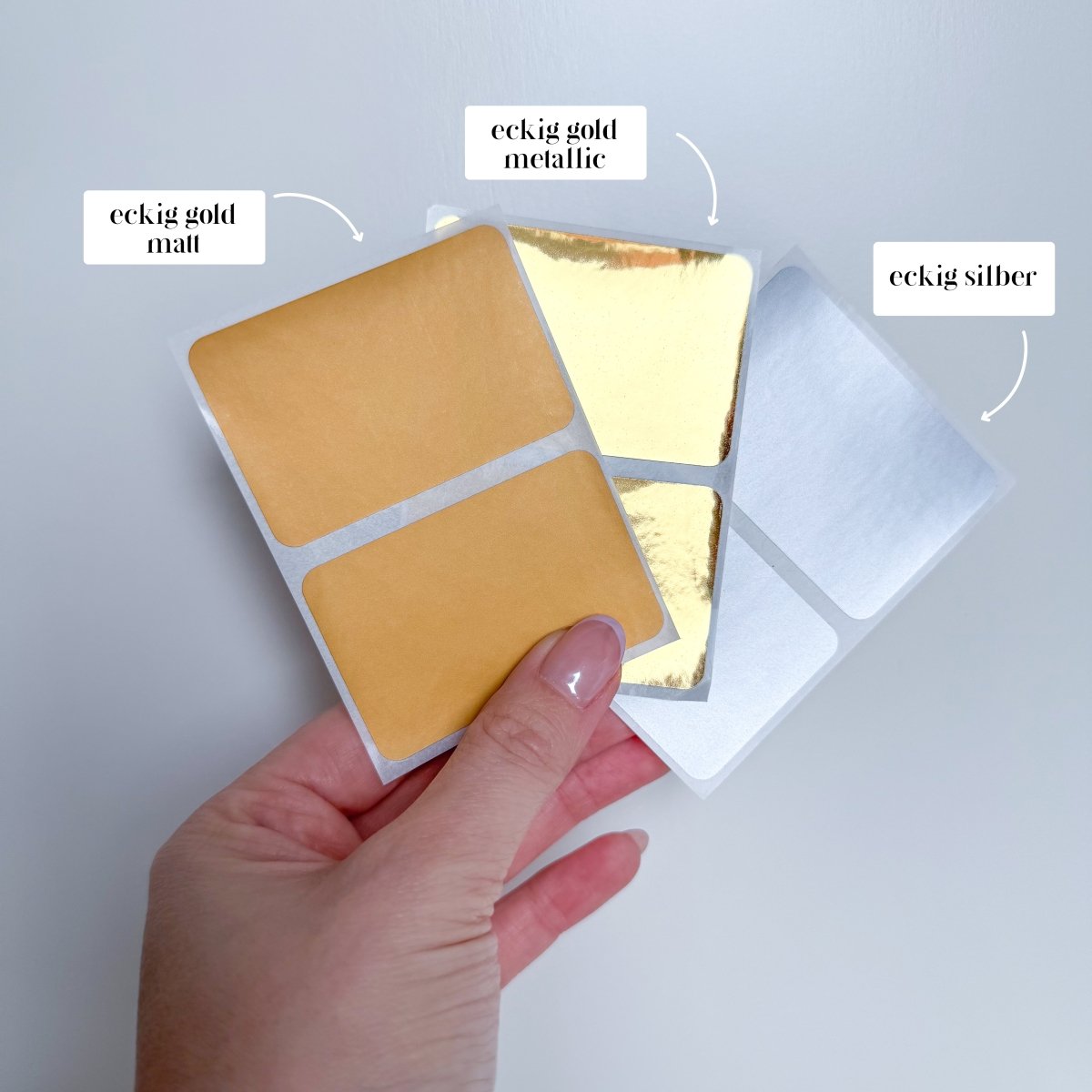 Eine Hand hält drei Bögen mit quadratischen Aufklebern auf weißem Hintergrund. Die Aufkleber sind in verschiedenen Ausführungen erhältlich: Mattgold, Metallicgold und Silber. Mit dem JGA-Reisegutschein „Personalisiert mit Umschlag“ von funkenschauer verleihen Sie jeder JGA-Feier eine persönliche Note.
