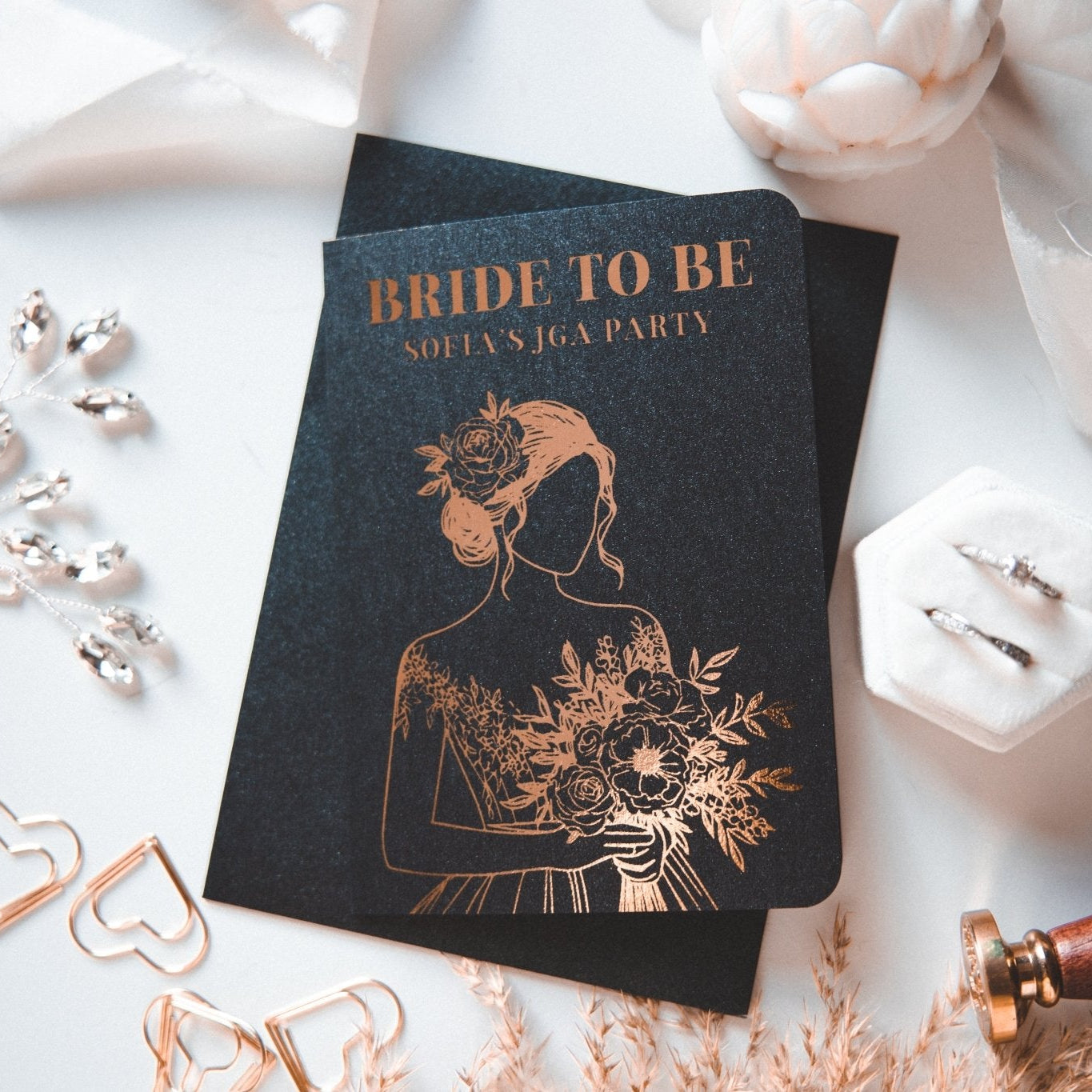 Elegante Einladung zur Brautparty auf schwarzem Karton mit einer Brautillustration in Goldfolie. Der Text lautet: „Zukünftige Braut, Sofias JGA-Party.“ Inklusive dekorativer Herzclips, Schmuck, Pampasgras und JGA-Frau-Ideen von funkenschauer – personalisierte Einladung mit Umschlag für Brautpartys.