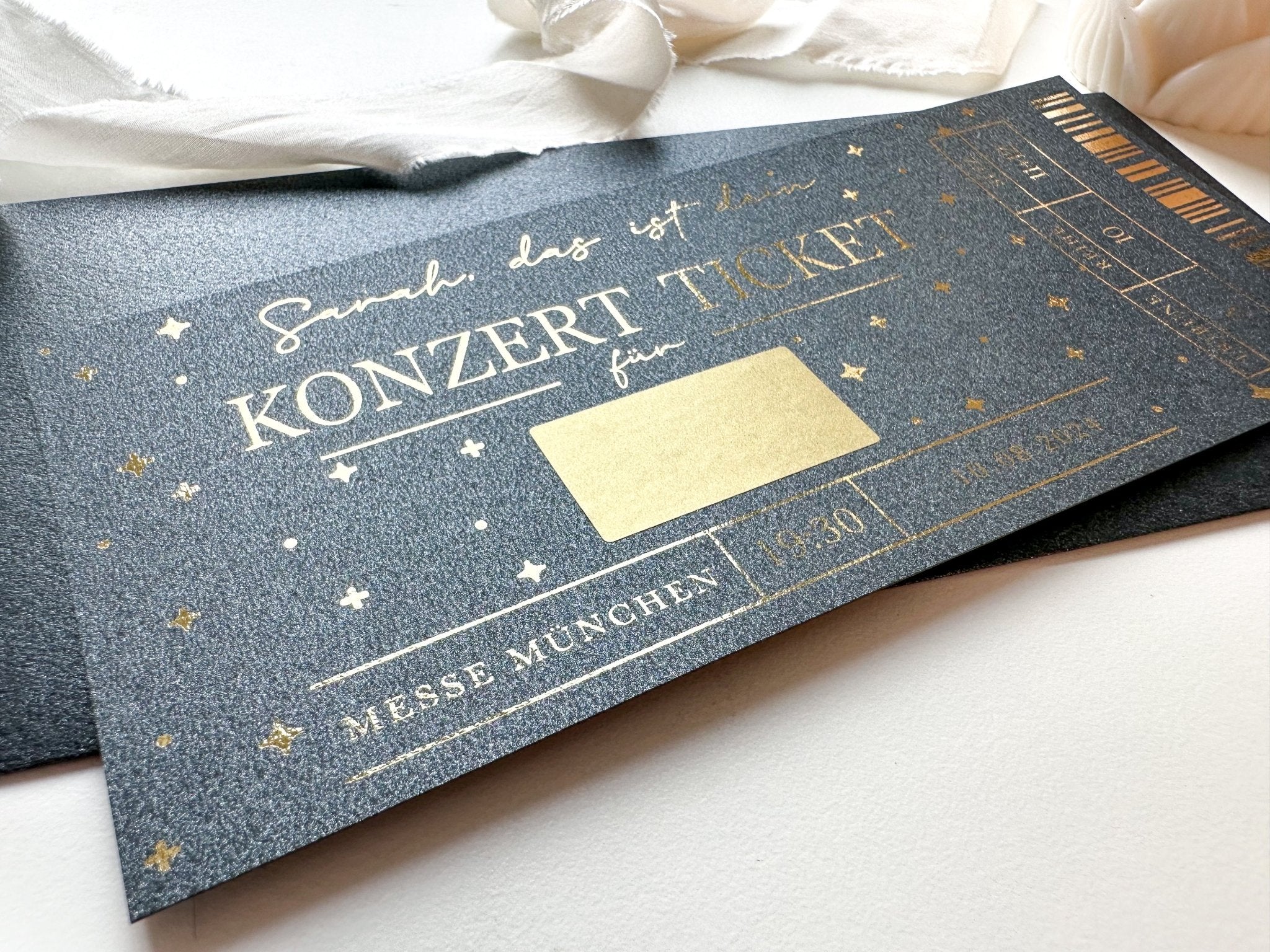 Eine Nahaufnahme eines Funkenschauer Gutschein Konzerts – eine personalisierbare Rubbelkarte für Musikliebhaber – mit goldener Schrift und Sternen auf schwarzem Papier, teilweise mit weißem Stoff bedeckt, „KONZERT TICKET“ und „MESSE MÜNCHEN“ gut sichtbar.