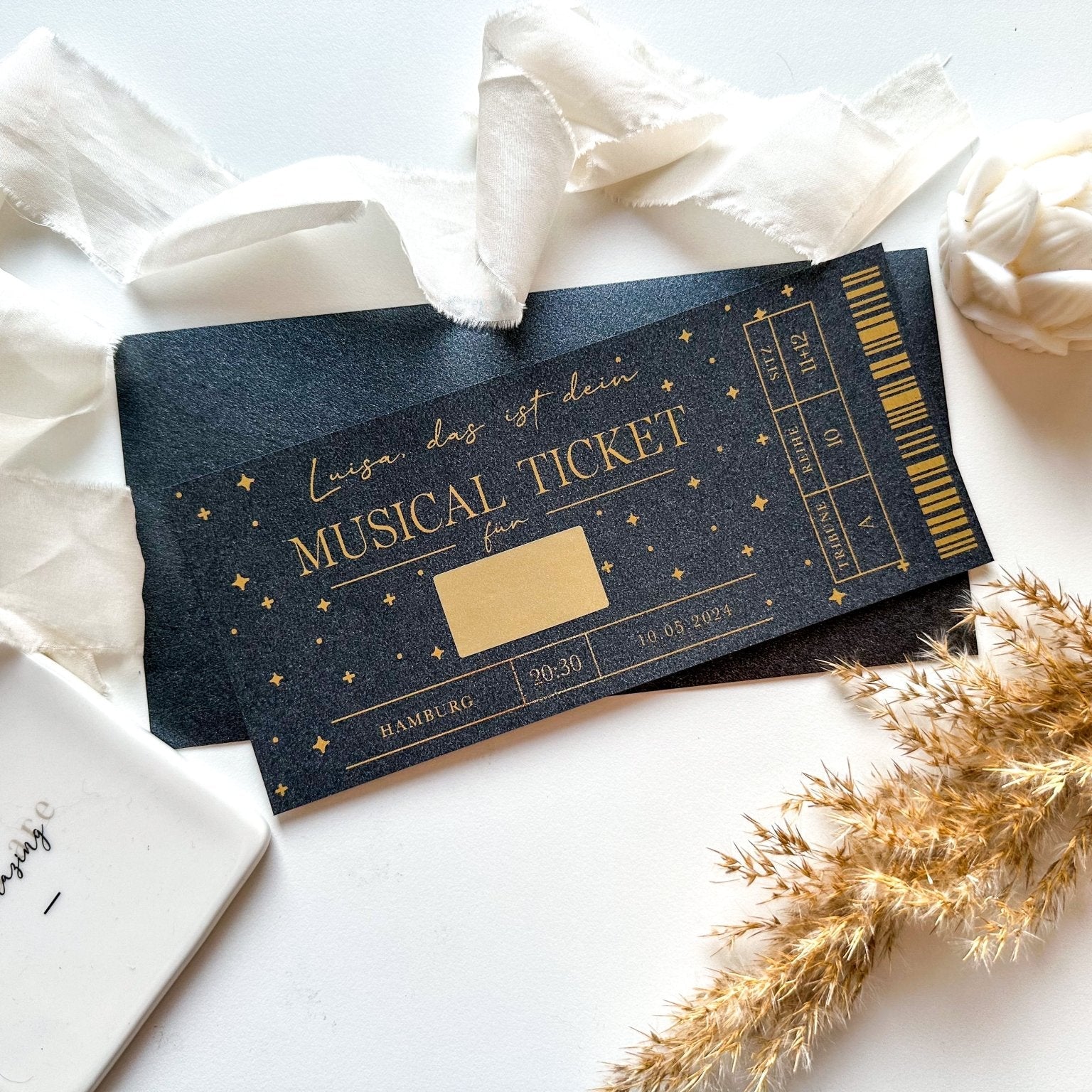 Der funkenschauer Musical-Gutschein • BLACK EDITION ist eine individuell gestaltbare Eintrittskarte für Shows wie „Der König der Löwen“ oder „Tarzan“, gestaltet mit Sternen, Text und Barcode – ein einzigartiges Geschenk, überreicht mit Schleife, getrocknetem Gras und einem Marmorblock.