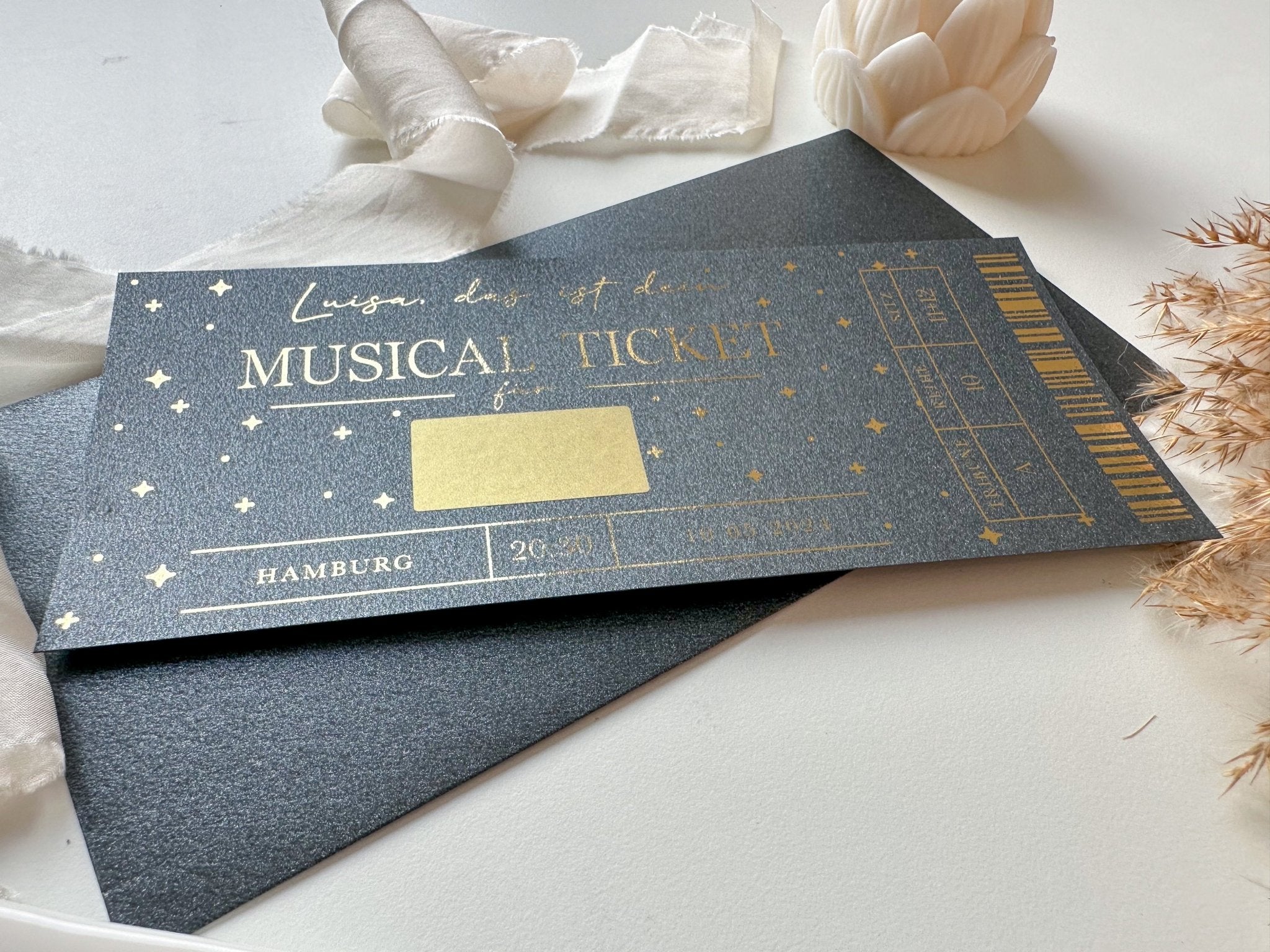 Ein funkenschauer Musical Gutschein • Ticket Personalisierbar in BLACK EDITION, verziert mit Sternen und elegantem Text, liegt auf einem weißen Umschlag – das perfekte Rubbelkarte Eventgutschein für Musicals wie Der König der Löwen oder Tarzan.