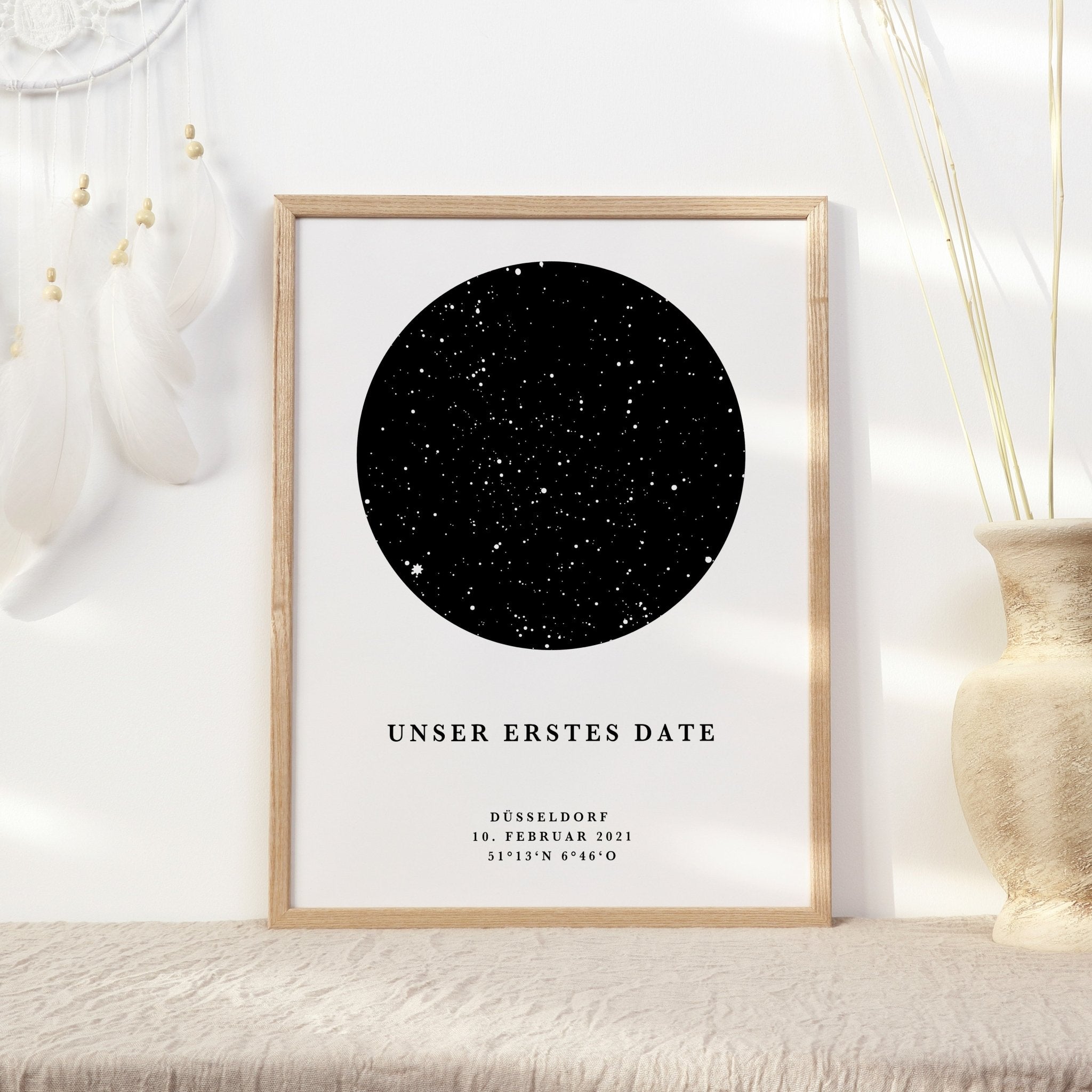 Gerahmter Druck mit einem „Sternenhimmel Poster personalisiert“ von funkenschauer mit dem Titel „Unser erstes Date“ und den Angaben: „Düsseldorf, 10. Februar 2021“ und Koordinaten. Es ist schwarz umrandet und mit Federn und einer Vase geschmückt.