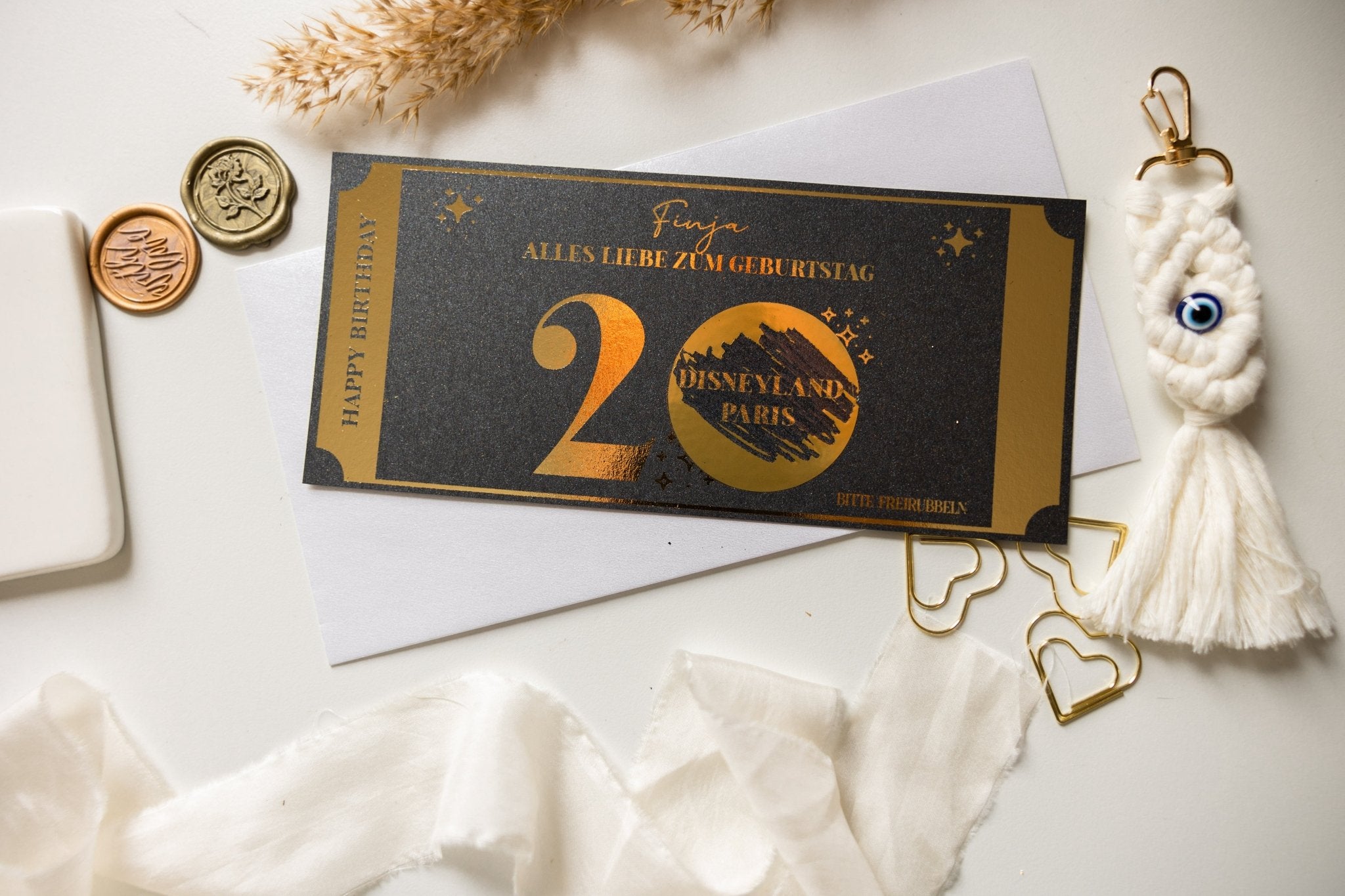 Ein funkenschauer personalisierter Geburtstagsgutschein mit Rubbelfeld für einen runden Geburtstag zeigt ein elegantes Design mit „Disneyland Paris“ und „20“ in Gold. Er ist umgeben von einem Wachssiegel, Schlüsseln, Pampasgras und einem Makramee-Schlüsselanhänger auf einer hellen Oberfläche auf einem weißen Umschlag.