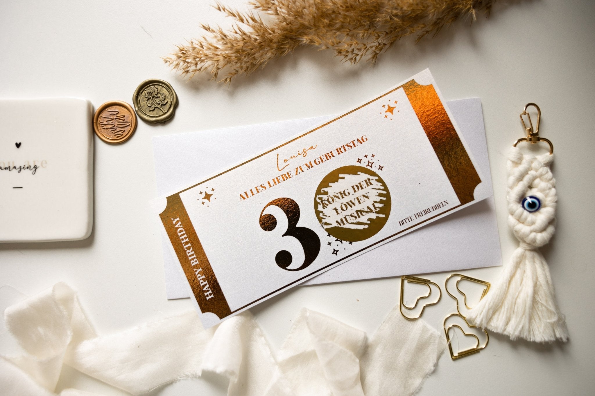 Ein funkenschauer Geburtstagsgutschein mit Edles-Design und goldenen Akzenten zum 30. Geburtstag liegt auf einer weißen Oberfläche, begleitet von goldenen Herzklammern, Pampasgras, einer Augen-Charm-Quaste und einem mit Wachs versiegelten Buchstaben. Der personalisierte Erlebnisgutschein passt zu jedem Stil.