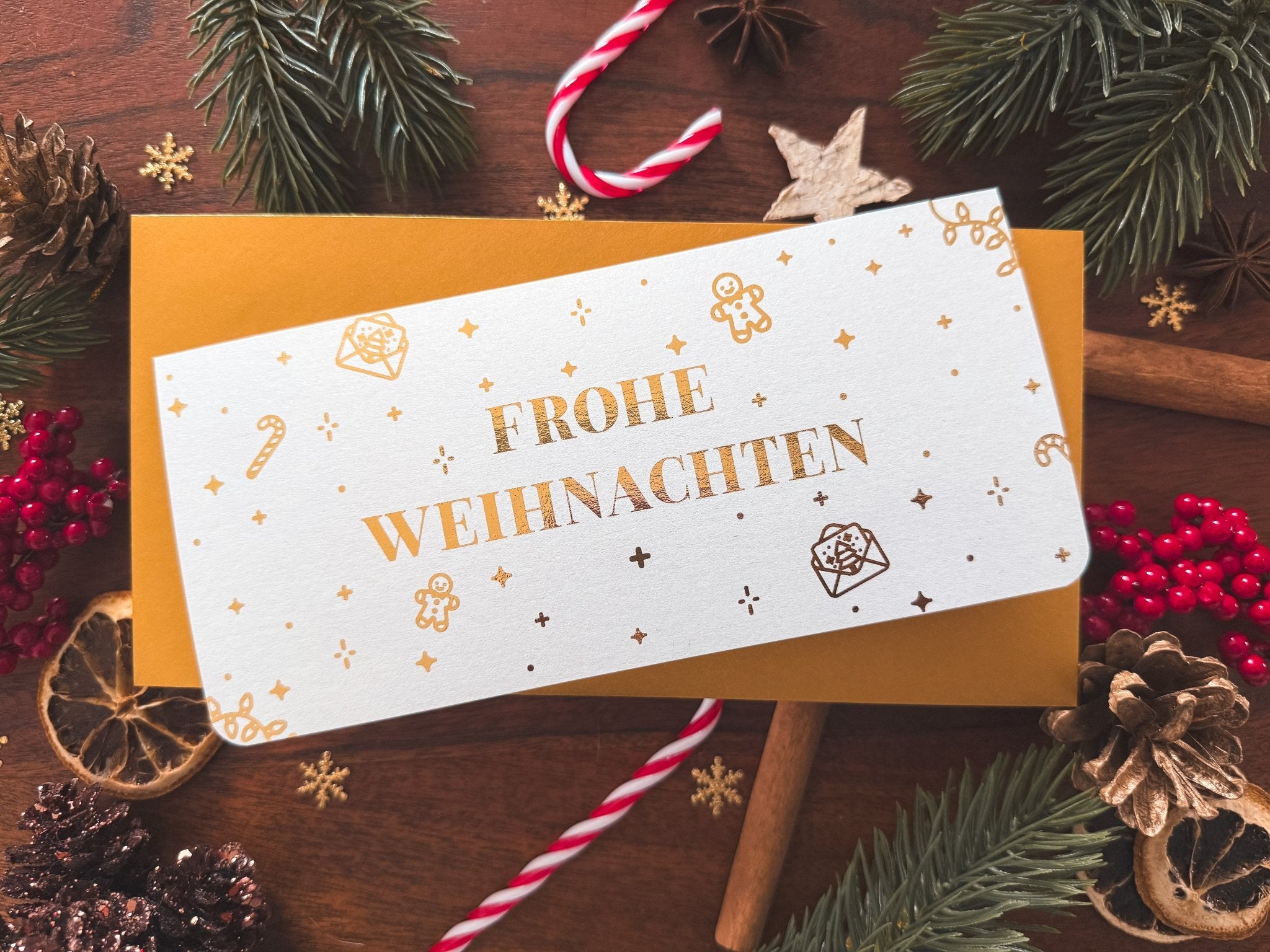 Die funkenschauer "Originelle Weihnachtsgeschenke" personalisierte Rubbelkarte "Frohe Weihnachten" bietet Zeit statt Zeug und liegt auf einem goldenen Umschlag - umgeben von Tannenzweigen, Beeren, Zimt und getrockneten Zitrusfrüchten.