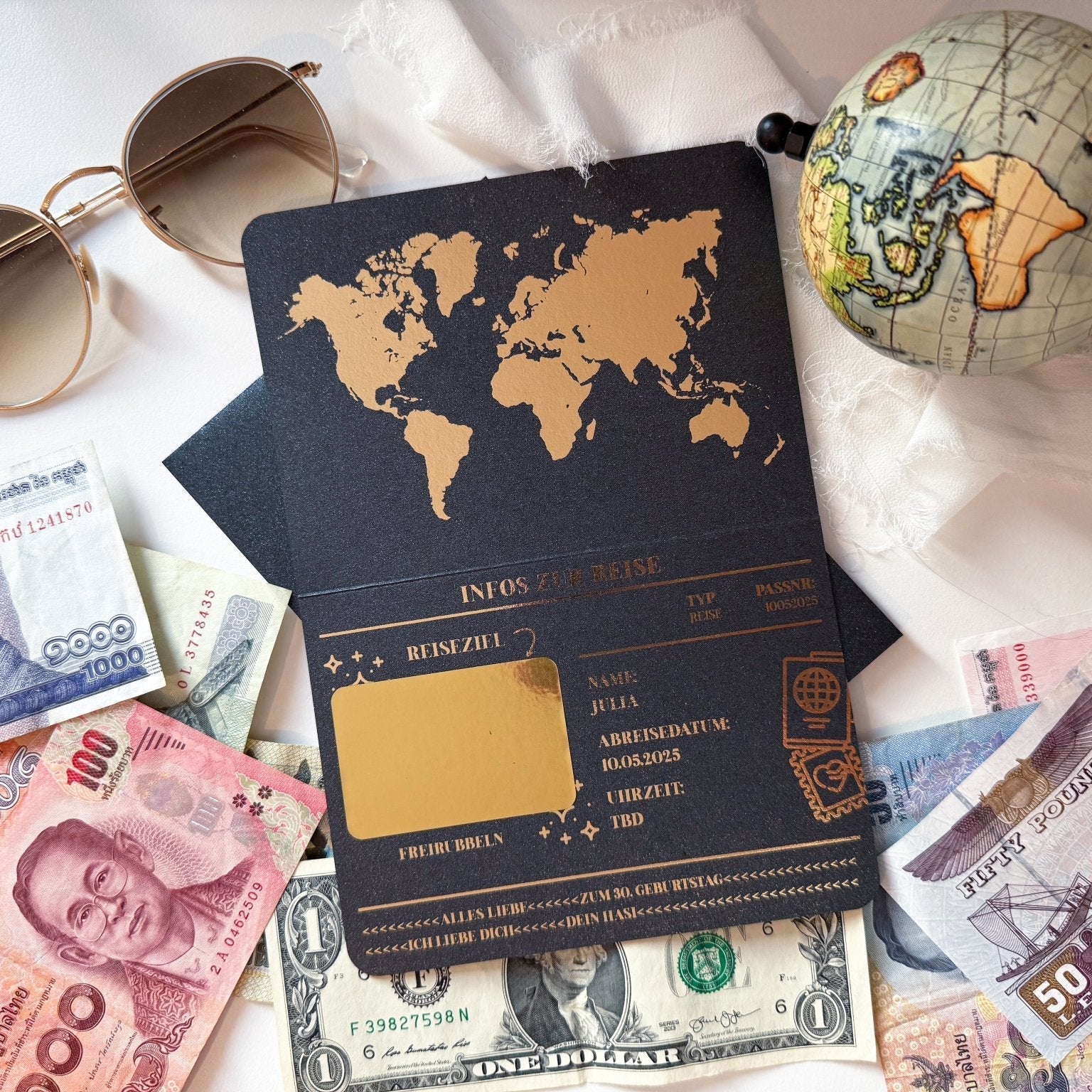 Flachgelegte Reiseartikel: Sonnenbrille, kleiner Globus, diverse Banknoten (US-Dollar und asiatische Währungen) und eine schwarze Mappe mit dem funkenschauer-Urlaubs-Verschenken-Pass im Weltkarten-Design und Rubbelkarte für personalisierte Abenteuer-Geschenke.