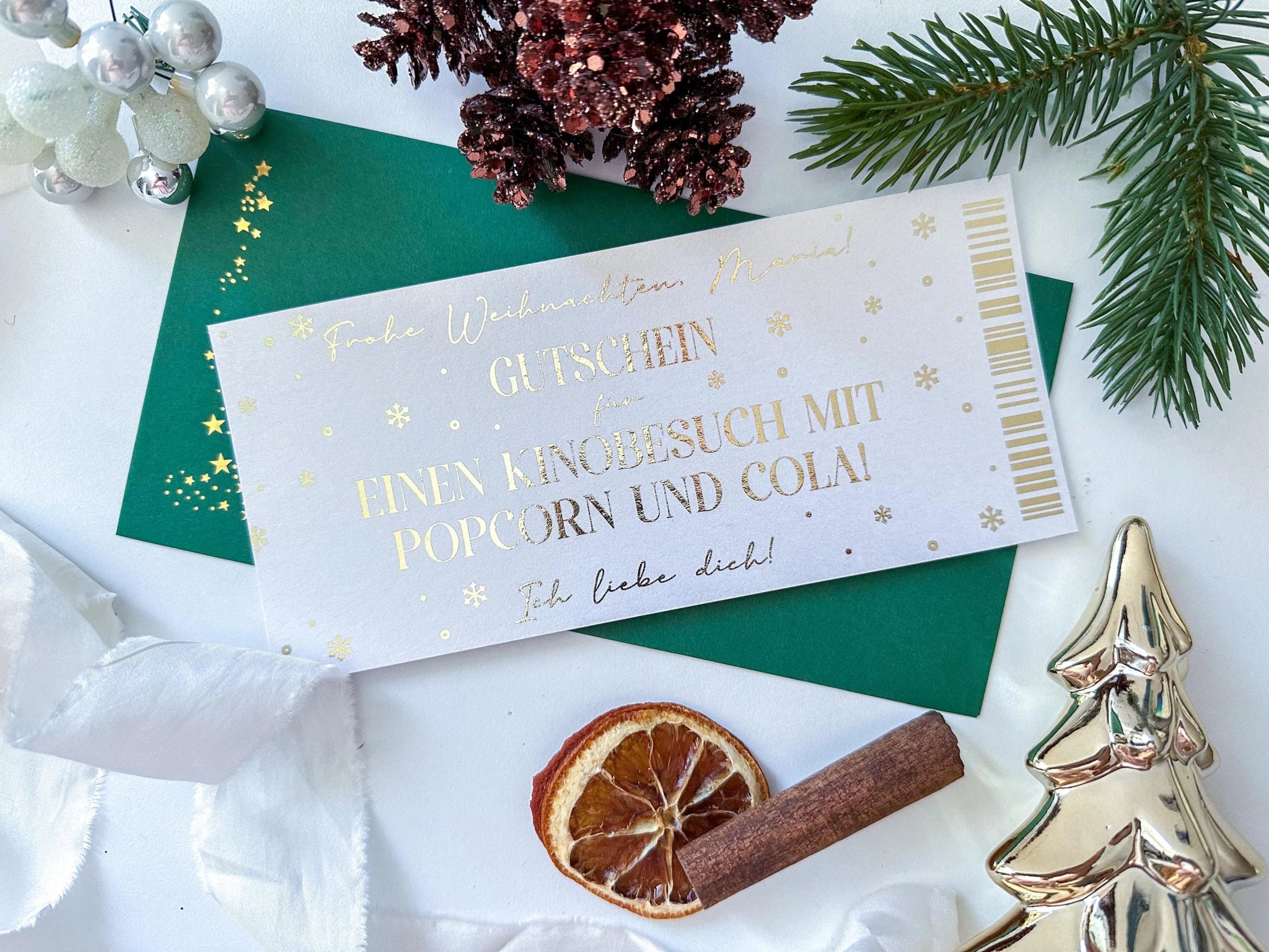 A HANDGEMACHTE WEIHNACHTSGESCHENKE PERSONALISIERTER WEIHNACHTSGUTSCHEIN von funkenschauer liegt auf einem weiΓen Tisch mit Tannenzweigen, Deko und Umschlag. Der Gutschein verspricht: "Kinobesuch mit Popcorn und Cola!.