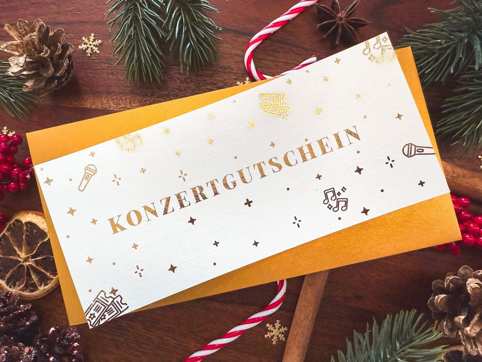 Auf einem Holztisch mit Tannenzapfen, immergrünen Zweigen, roten Beeren, Zuckerstangen und getrockneten Orangenscheiben liegt ein „Konzertgutschein Personalisiert“ von Funkenschauer mit der Aufschrift „KONZERTGUTSCHEIN“ in goldenen Buchstaben; Dieses luxuriöse Ticketdesign verspricht unvergessliche Momente für Musikliebhaber.