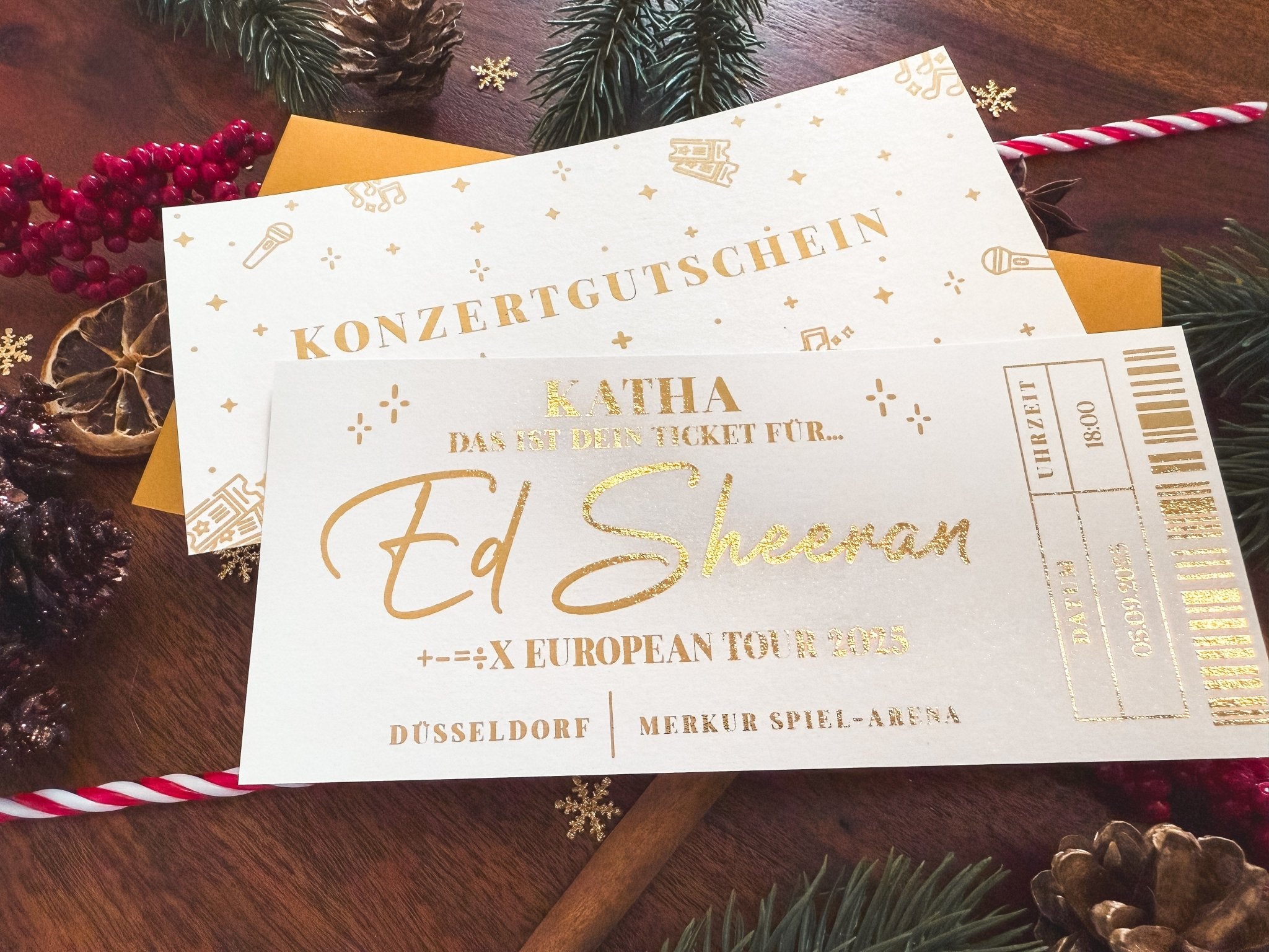 Auf einem festlich dekorierten Holztisch liegt ein luxuriöser Konzertgutschein von Funkenschauer. Dieses exquisit gestaltete Ticket bietet ein personalisiertes Erlebnis-Ticket für ein Ed Sheeran-Konzert in der Düsseldorfer Merkur Spiel-Arena, Teil der +-=÷× European Tour 2023.