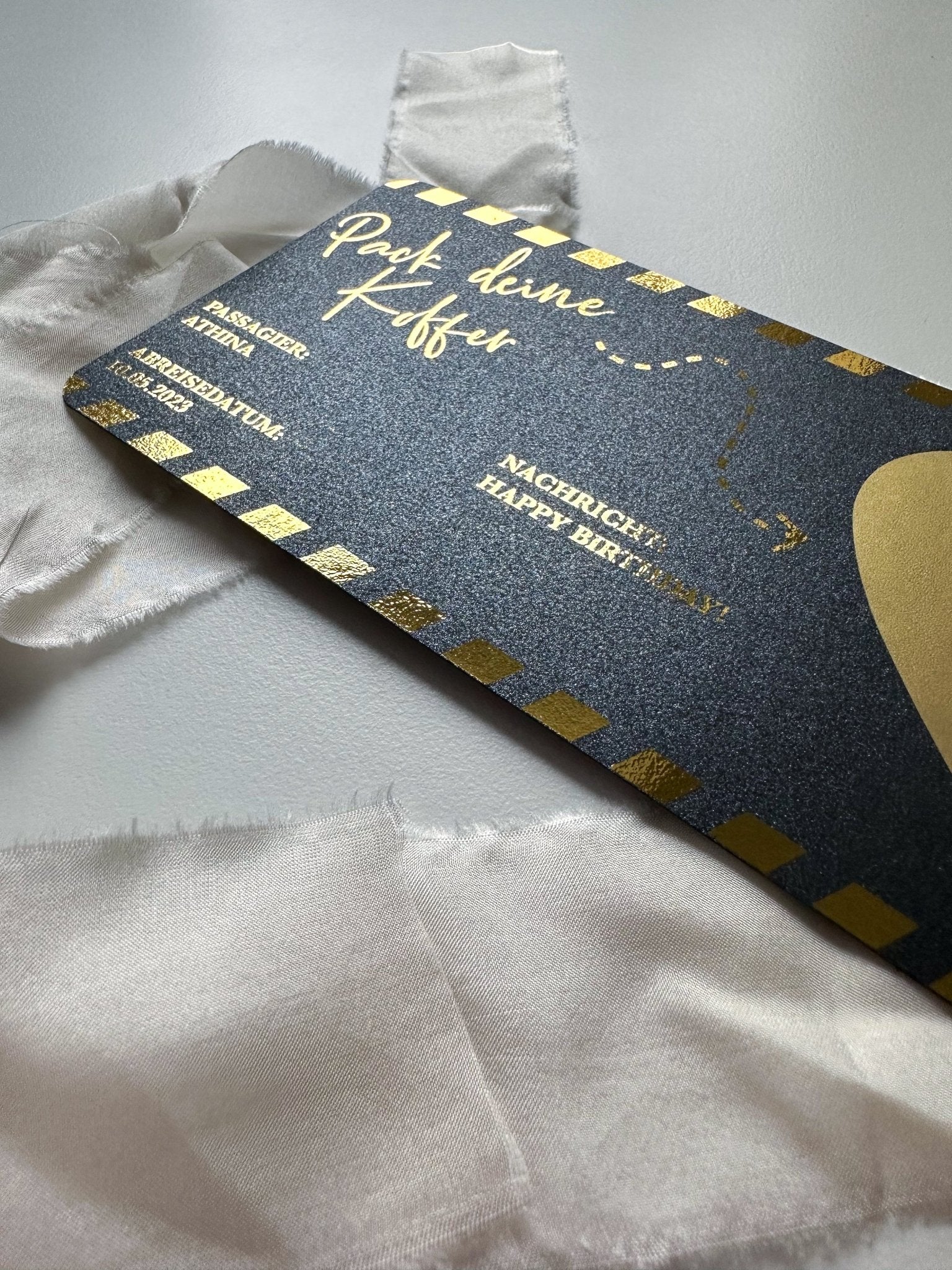 Eine dunkle Reisegutschein-Verschenken-Karte von Funkenschauer mit goldenen Akzenten und einer Schleife liegt auf einer weißen Oberfläche. Die Botschaft „Pack deinen Koffer“ deutet auf ein abenteuerliches Geschenk hin, das sich in dieser personalisierbaren Überraschung verbirgt.