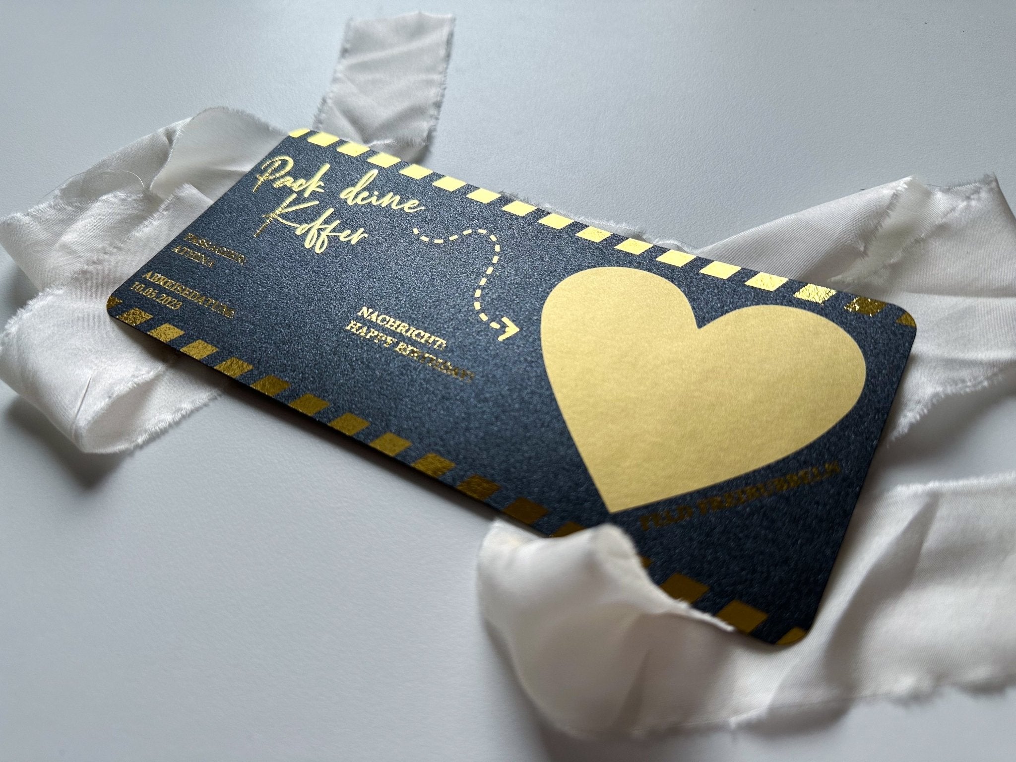 Ein funkenschauer Reisegutschein mit goldener Schrift und einem großen Herzen auf dunklem Hintergrund auf weißem Grund. Die Ränder der Karte sind mit goldenen Streifen versehen, die auf ein aufregendes Geschenk im Inneren hinweisen. Ein weißes Band umschließt diesen personalisierbaren Schatz für Reiseliebhaber elegant.