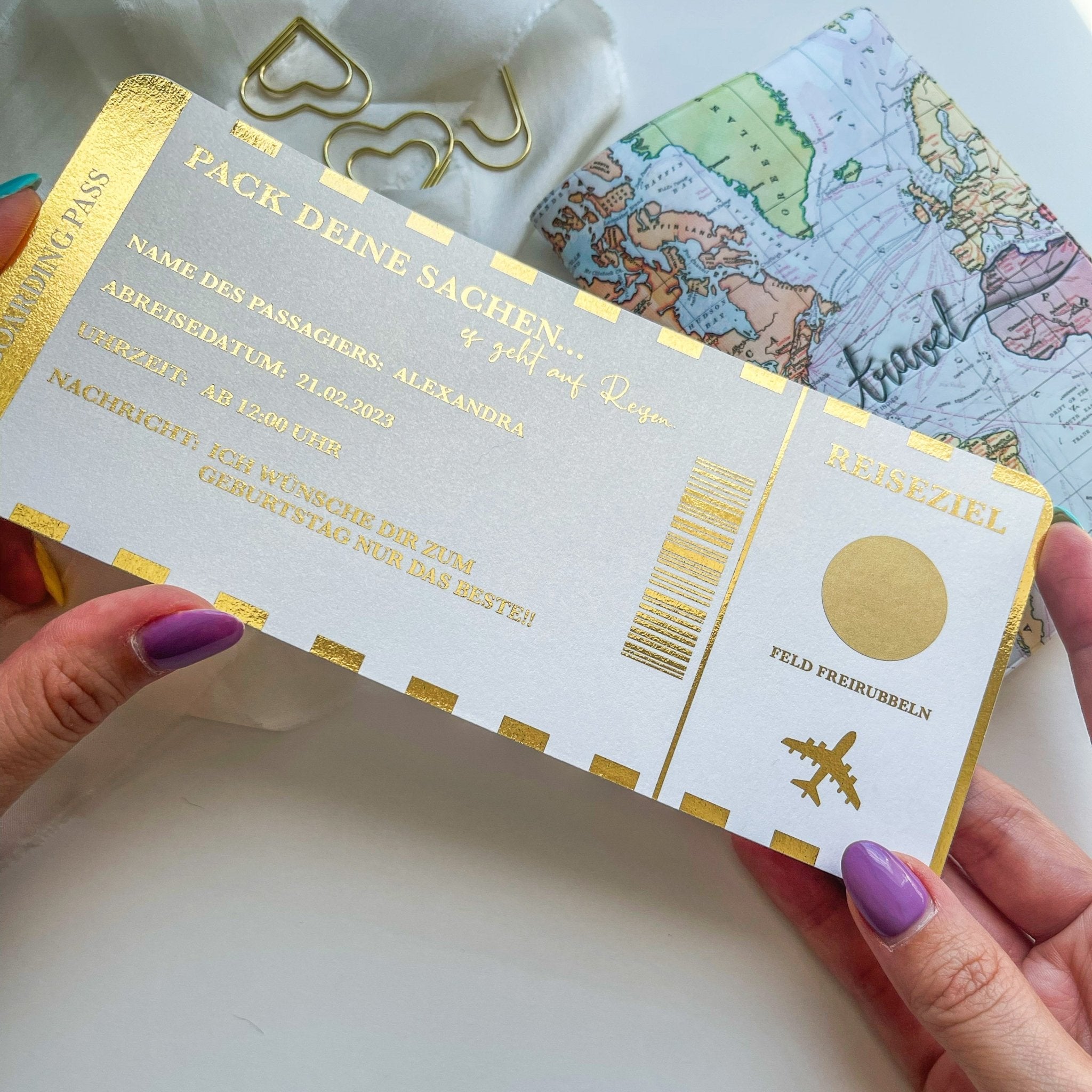 Jemand hält einen gold-weißen Funkenschauer „Reisegutscheine zum Verschenken“ im Bordkarten-Stil mit deutschem Text neben einem Reisenotizbuch, herzförmigen Büroklammern und einer Karte – ein perfekter personalisierter Reisegutschein für Weltenbummler.