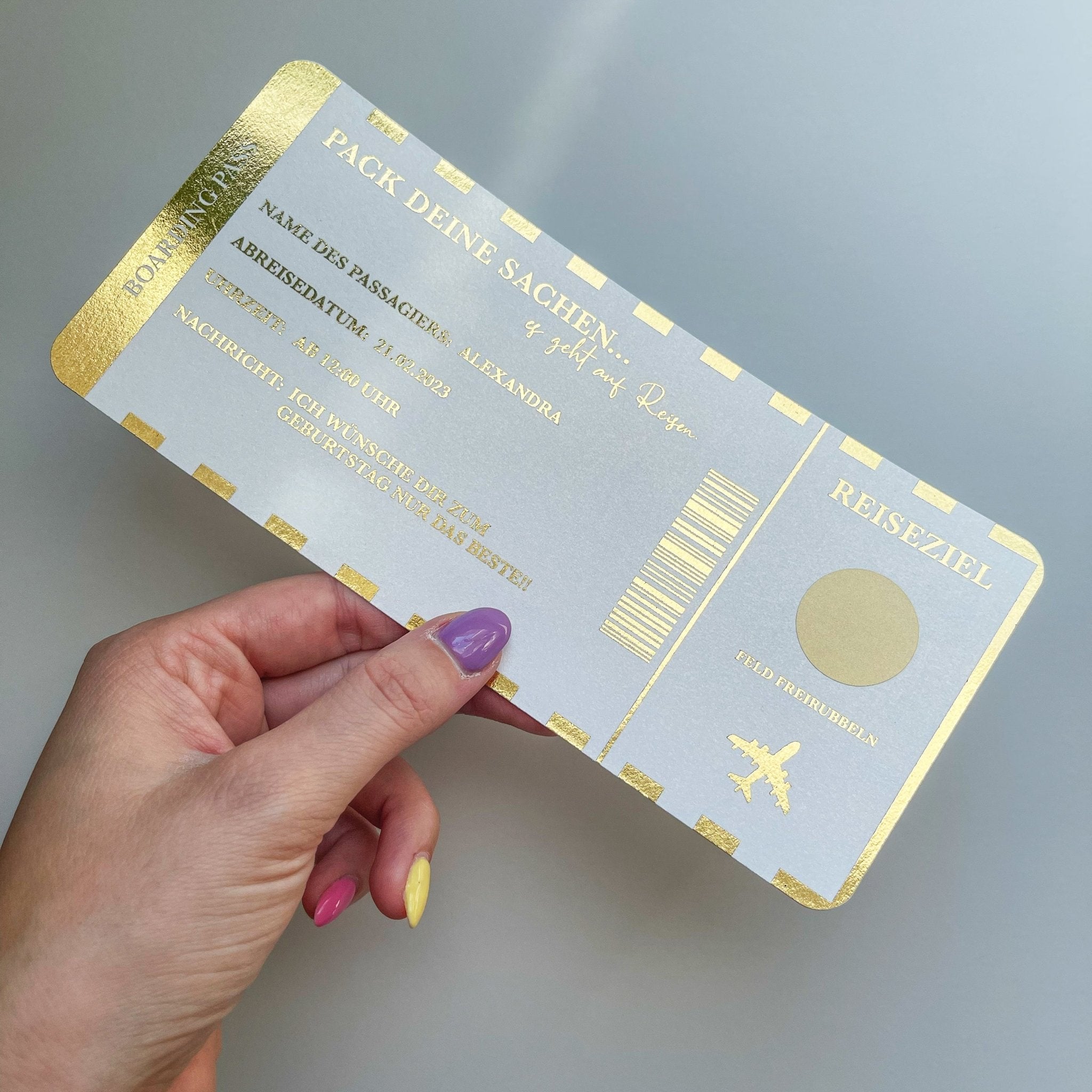 Eine Hand mit bunten Nägeln hält einen gold-weißen Funkenschauer Reisegutscheine zum Verschenken im Bordkarten-Stil mit Rubbelfeld – das perfekte Geschenk für Reiseliebhaber und Weltenbummler – vor hellgrauem Hintergrund.