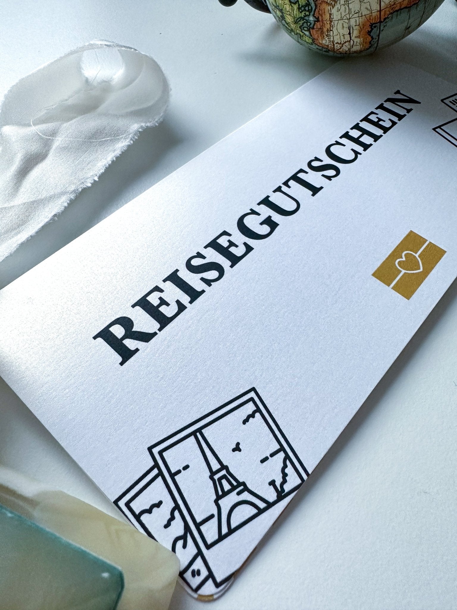 Auf einer weißen Unterlage liegt ein Funkenschauer-Reisegutschein mit Reisegutschein und bezaubernden Pariser Illustrationen, in der Ecke ein Globus. Diese personalisierte Geschenkidee ist perfekt für Fernweh-Enthusiasten.