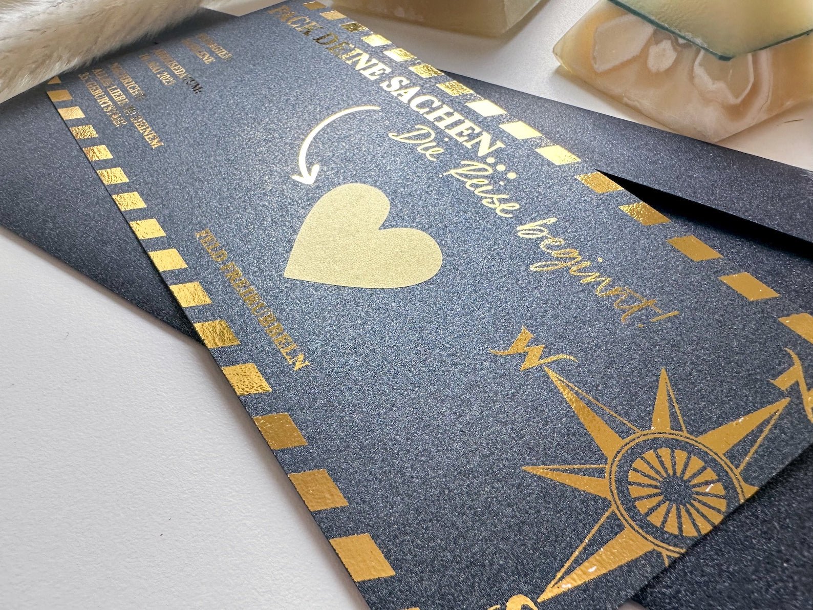 Eine Nahaufnahme eines dunklen, schimmernden Funkenschauer Reisegutscheins im Bordkarten-Design mit Goldfolie „Die Reise beginnt“, deutscher Text, gestreifter Rand, Herz, Windrose – ein ideales personalisiertes Geschenk mit Rubbelkarte für Reiseliebhaber.