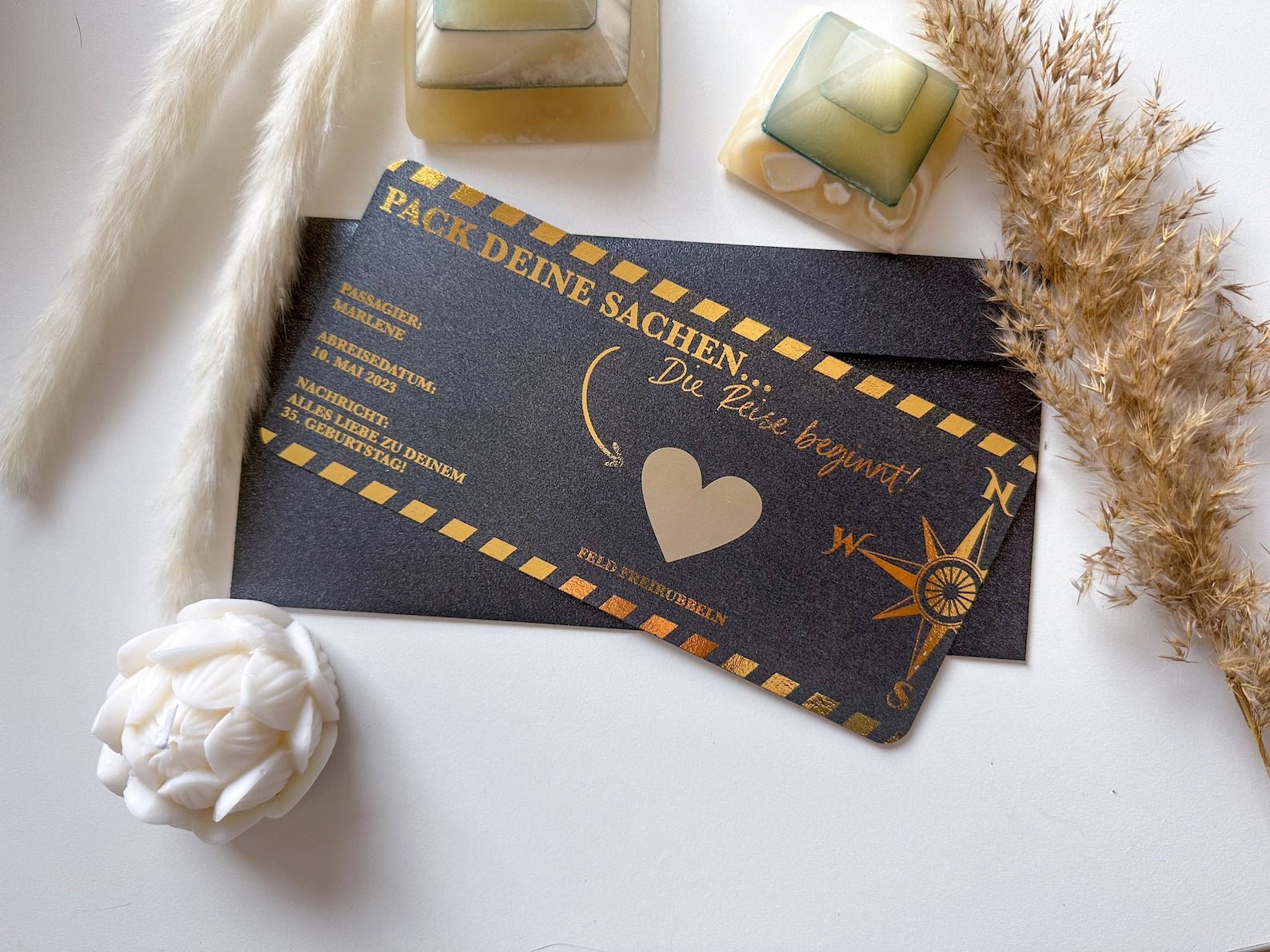 Zwei schwarze und goldene Funkenschauer Reisegutscheine als Bordkarte mit Rubbelfeld und Herz-Kompass-Motiv liegen auf weißem Untergrund, umgeben von Kerzen und Deko – das perfekte personalisierte Geschenk für Reiseliebhaber.