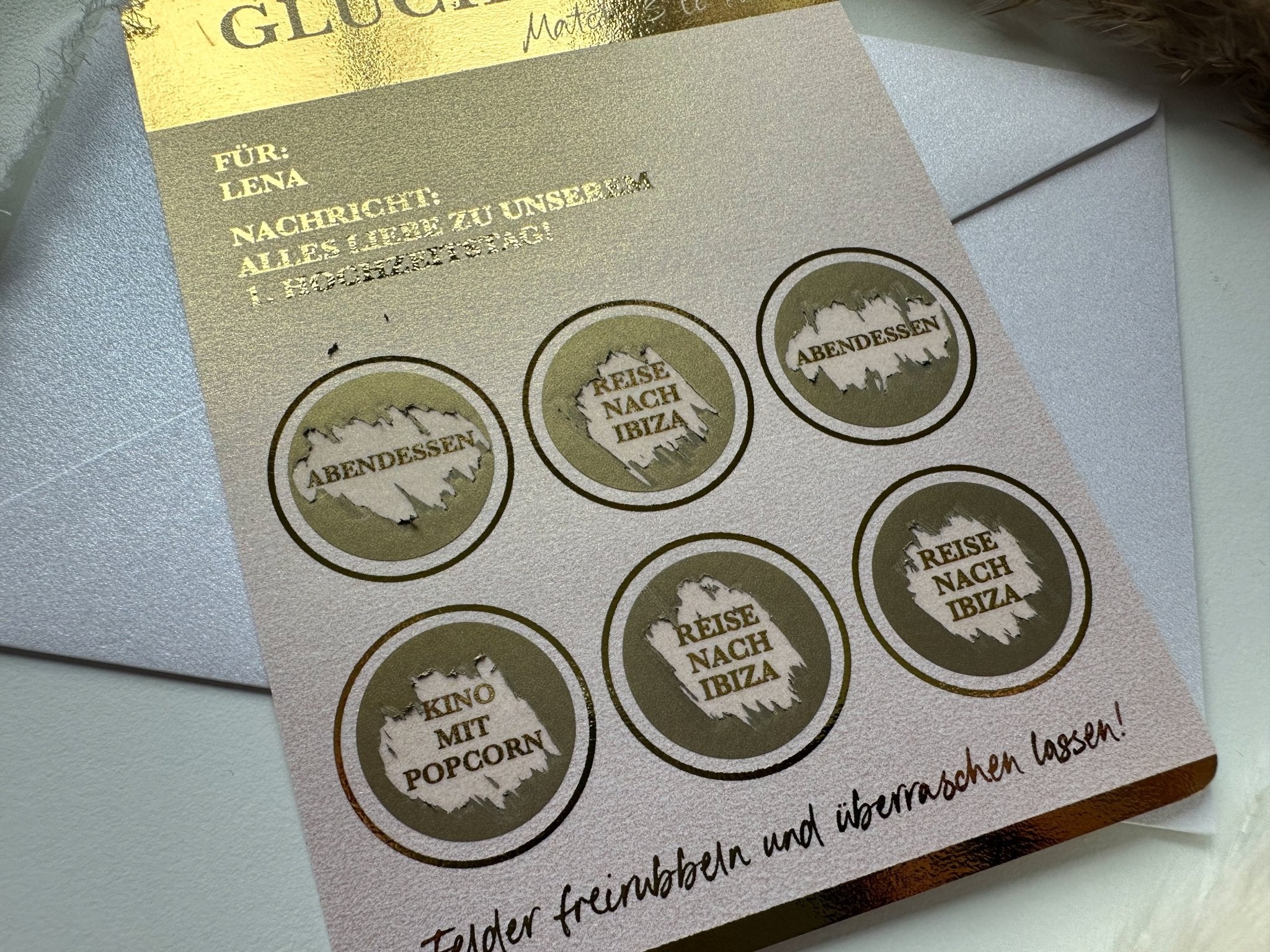 Eine Nahaufnahme des Personalisierbaren Rubbellos Inkl. Kuvert von funkenschauer zeigt sechs runde Felder, auf denen jeweils zwei deutsche Sprüche wie „Abendessen“, „Kino mit Popcorn“ und „Reise nach Ibiza“ stehen. Die Rubbelkarte hat einen metallisch-goldenen Rand und liegt auf einem weißen Umschlag, was sie zu einem idealen personalisierten Geschenk aus der Zeit statt Zeug-Kollektion macht.