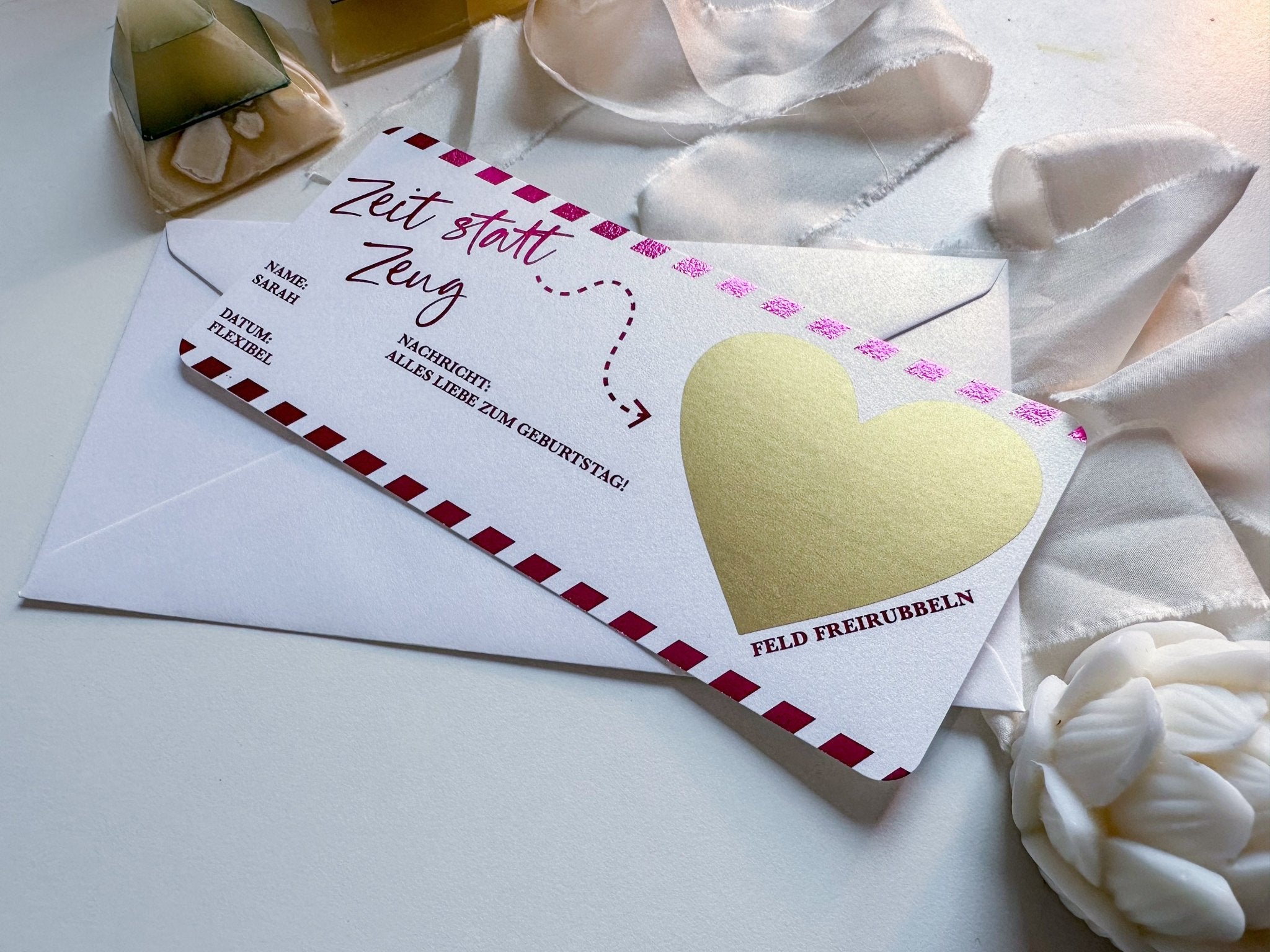 Auf einem weißen Umschlag liegt ein dekorativer „Zeit statt zeug Gutschein“ von Funkenschauer mit goldenem Herz und roten Streifen. Der deutsche Text deutet auf eine Geburtstagsnachricht hin. Ein weißes Band, eine Blume und Kerzen betonen die Szene, ideal für alle, die personalisierte Geschenke suchen.
