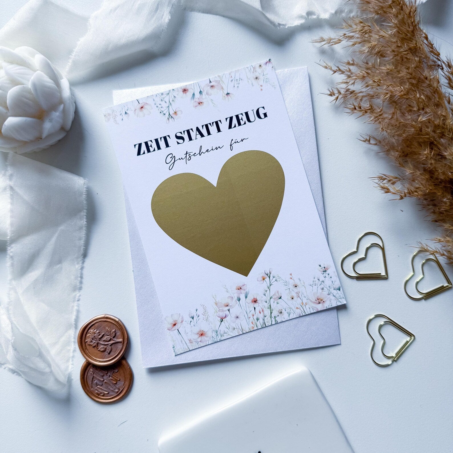 Ein „Zeit statt Zeug Gutschein – personalisiertes Geschenk“ von Funkenschauer, A6 Rubbelkarte mit floralem Design, großem Goldherz und deutschem Text liegt auf einem Umschlag umgeben von weißen Blumen, goldenen Herzclips und Münzen.
