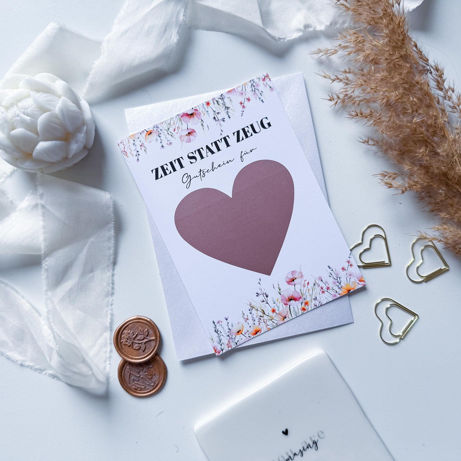 Die Funkenschauer Zeit statt Zeug Gutschein – personalisiertes Geschenk A6 Rubbelkarte verfügt über ein großes malvenfarbenes Herz, florale Akzente und wird mit dekorativem Band, weißer Blume, getrocknetem Gras, goldenen Clips und Wachssiegeln geliefert.