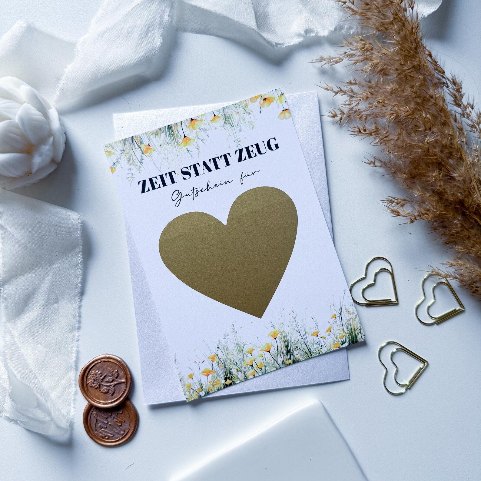 Die Funkenschauer Zeit statt Zeug Gutschein – personalisiertes Geschenk A6 Rubbelkarte liegt auf einem weißen Umschlag, umgeben von weißem Stoff, einer weißen Blume, goldenen Herz-Büroklammern, braunen Wachssiegeln und getrocknetem Gras.
