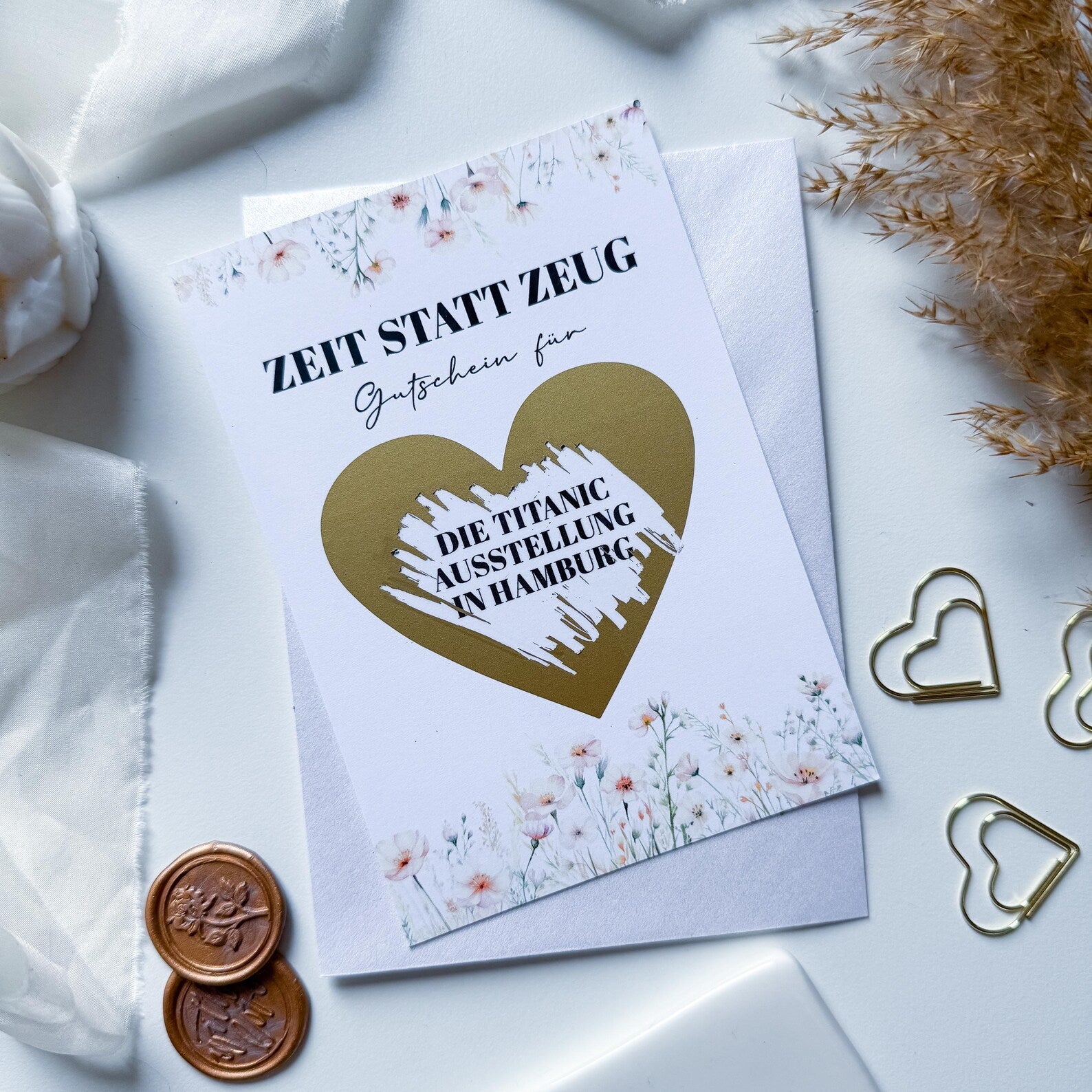 Eine florale „Zeit statt Zeug Gutschein – personalisiertes Geschenk A6 Rubbelkarte“ von Funkenschauer liegt auf einer weißen Fläche mit Münzen, Herz-Büroklammern und Akzenten. Es verfügt über ein goldenes Herz und Ihre persönliche Nachricht für gemeinsame Erlebnisse.