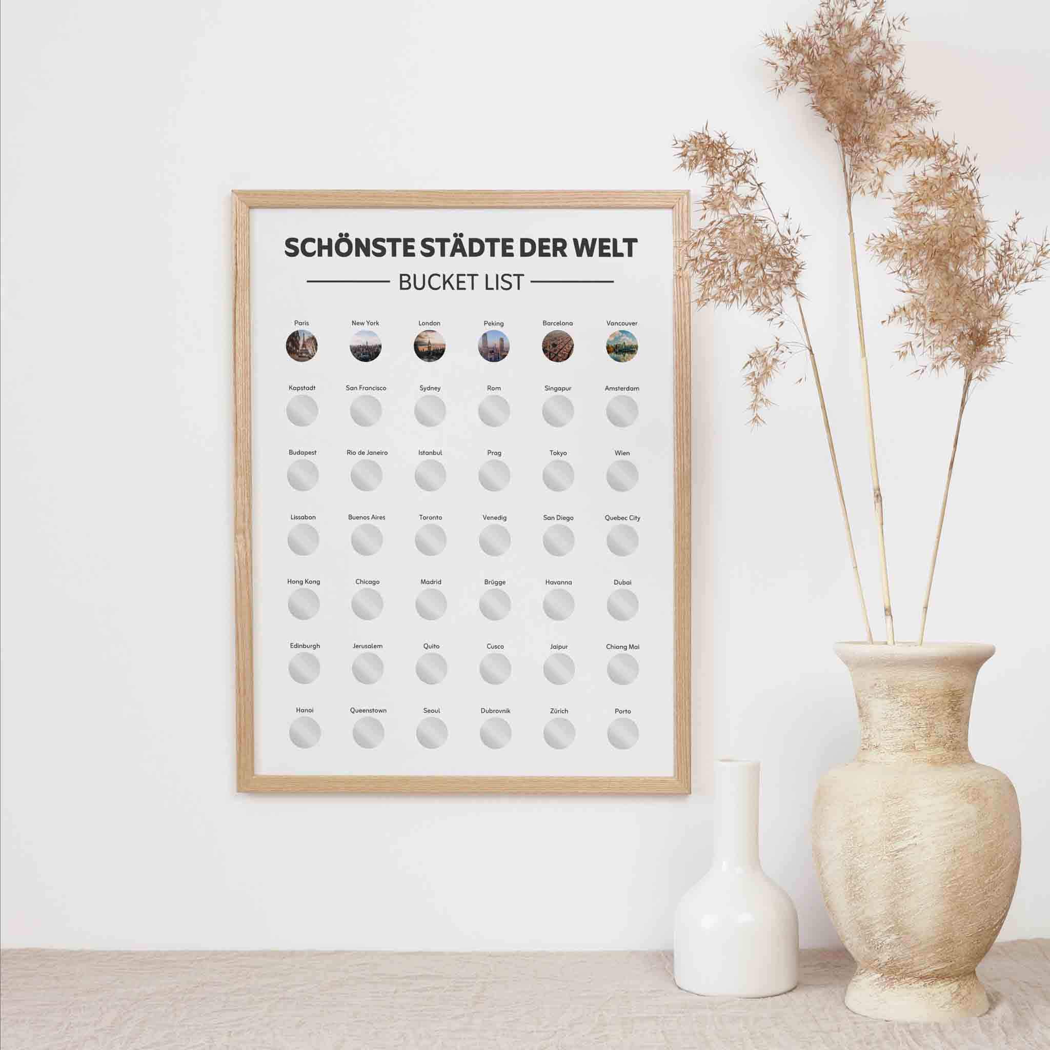 Das funkenschauer Poster "Schönste Städte der Welt Bucketlist zum Freirubbeln" (A3) mit runden Rubbelfeldern hängt an einer weißen Wand, daneben Trockenpflanzen in einer strukturierten Vase und eine weiße Keramikvase.