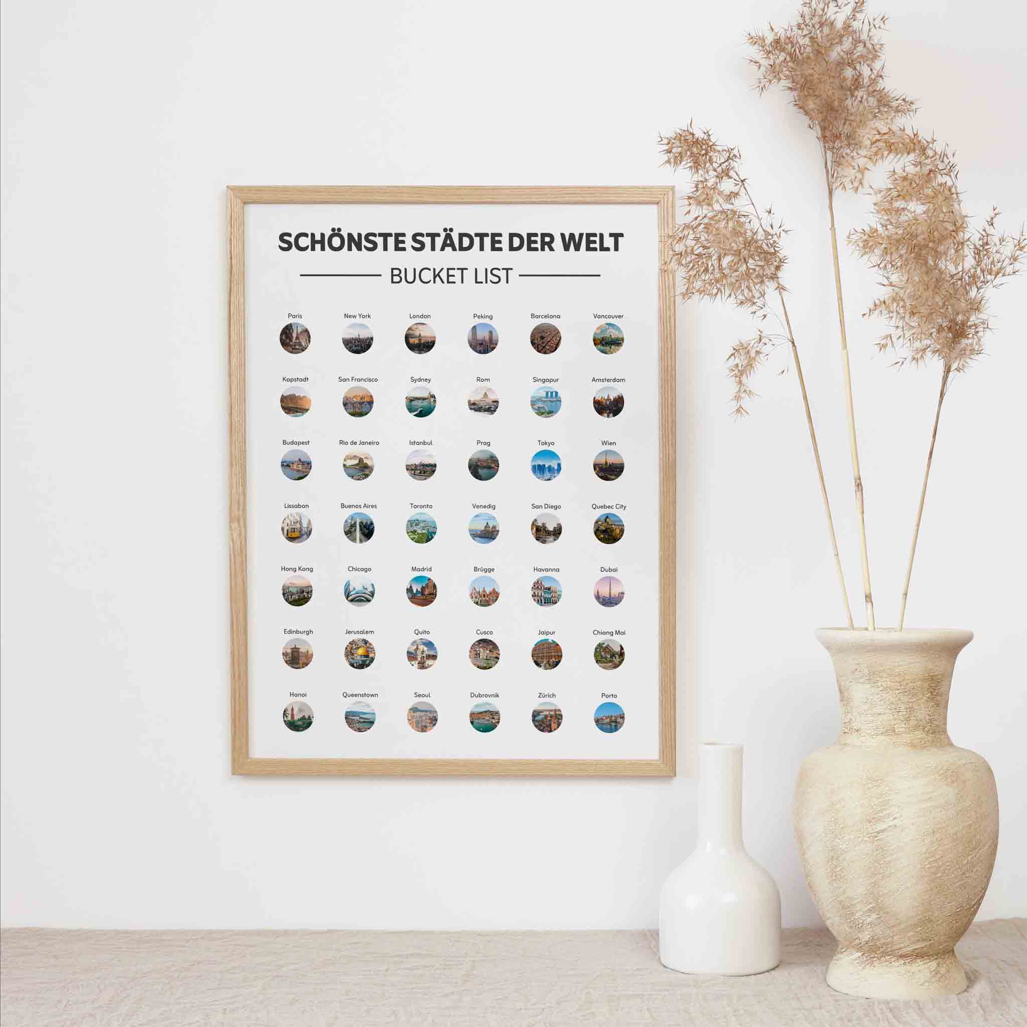 Das funkenschauer "Schönste Städte der Welt Bucketlist zum Freirubbeln" (A3) mit Fotos vieler Metropolen hängt neben einer beigen Vase mit Trockenpflanzen und einer weißen Keramikflasche an einer weißen Wand.