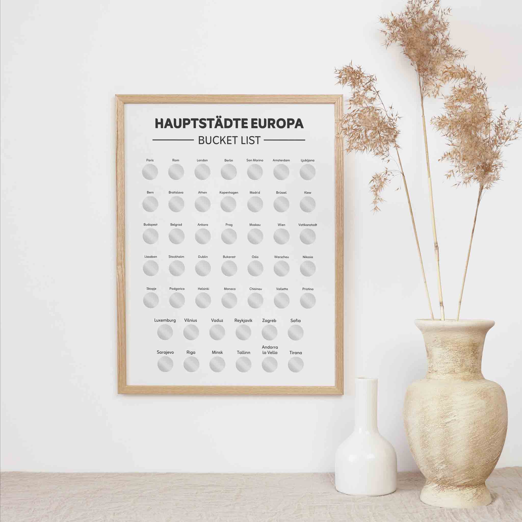 Das "Hauptstädte Europas Bucketlist zum Freirubbeln" Rubbelposter von funkenschauer hängt an einer weißen Wand neben getrockneten Gräsern in einer beigen Vase und zwei kleinen weißen Vasen.