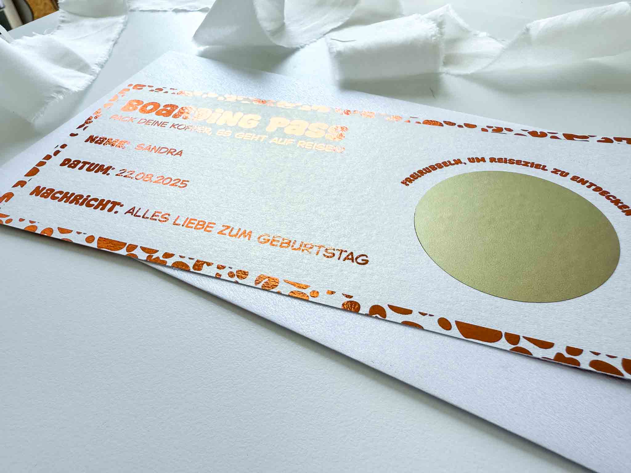 Eine Nahaufnahme von funkenschauers personalisiertem Reisegutschein Geburtstag Leoprint als Rubbelkarte im Boarding-Pass-Stil mit Namen, Datum und leuchtend orangefarbenen Akzenten, liegend auf Weiß mit einem Band im Hintergrund.