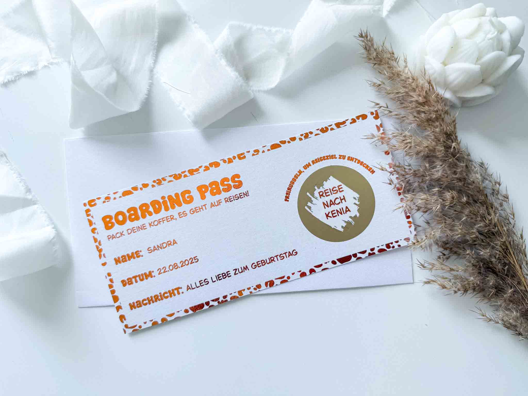 Ein funkenschauer Reisegutschein Geburtstag Leoprint als personalisiertes Reiseticket liegt auf einem weißen Umschlag, begleitet von Trockenblumen und Band. Die Rubbelkarte bietet Felder für Name, Datum und Geburtstagsbotschaft für eine Kenia-Reise.