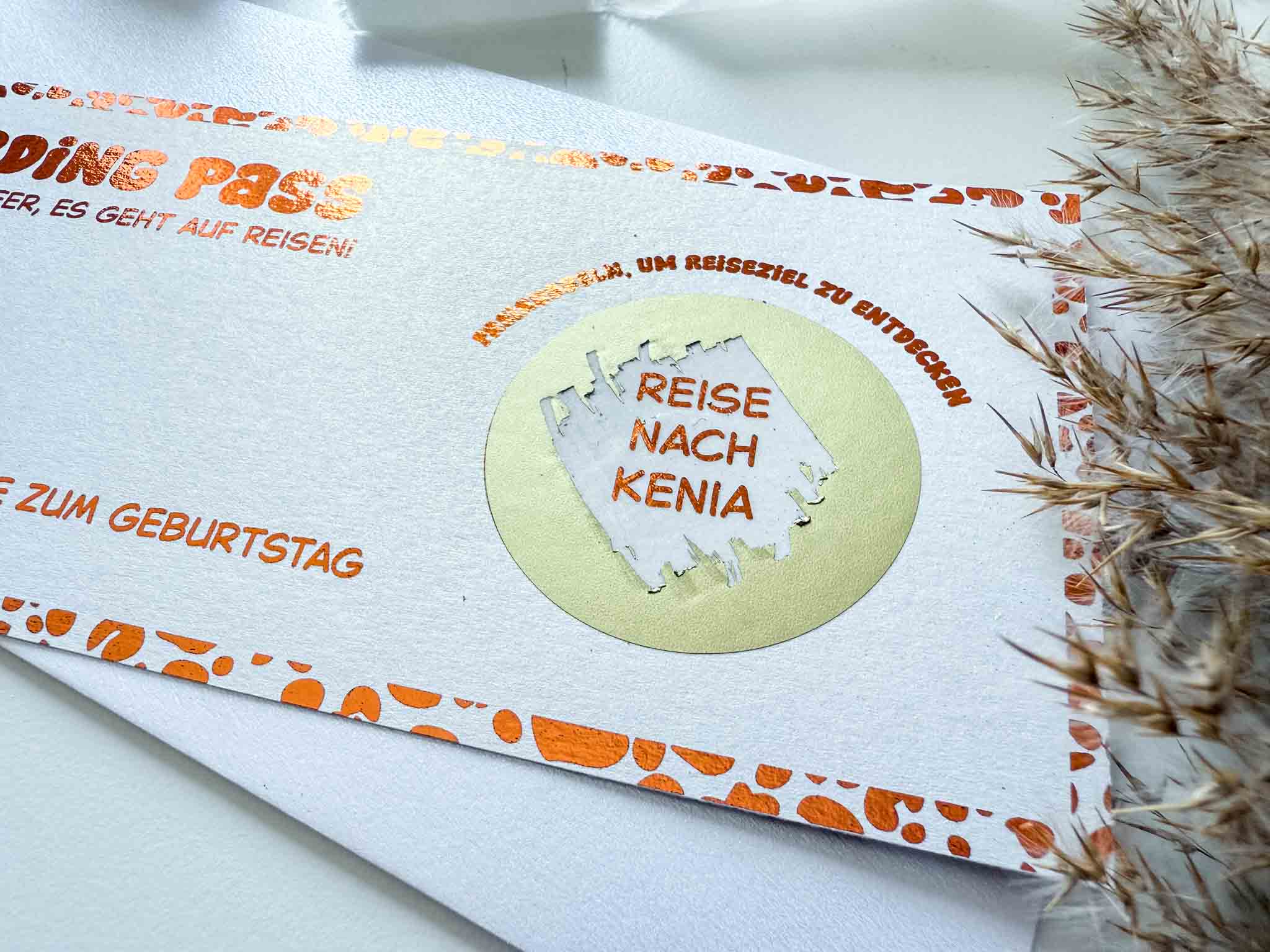 Eine Nahaufnahme des funkenschauer Reisegutscheins Geburtstag Leoprint Personalisiert Rubbelkarte im Boarding-Pass-Design mit Rubbelfeld "Reise nach Kenia", weißem Umschlag darunter und getrockneten Gräsern rechts.